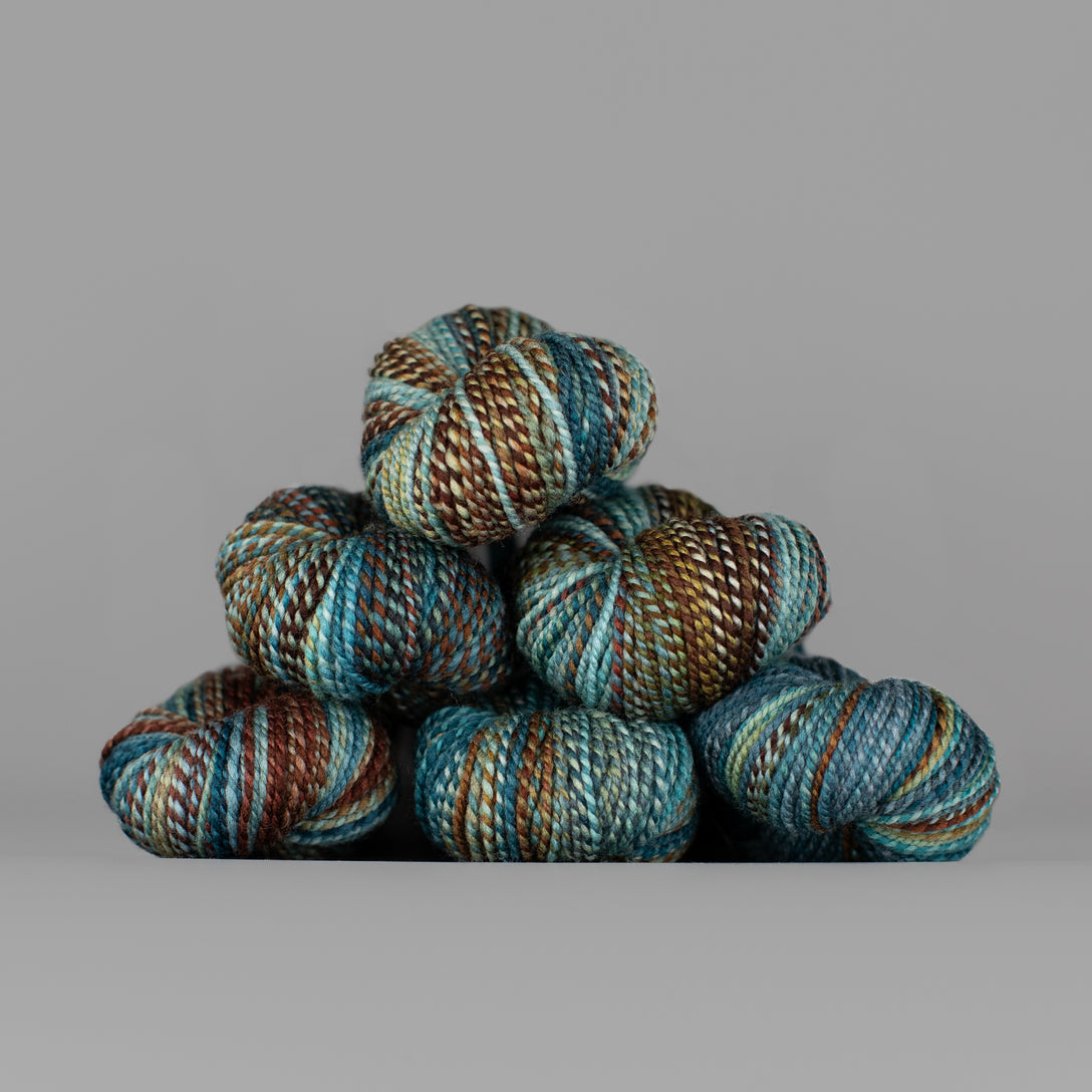 Skeins of Spincycle Dyed in the Wool sport weight yarn in a pale blue, teal, and brown color gradient against a gray background.