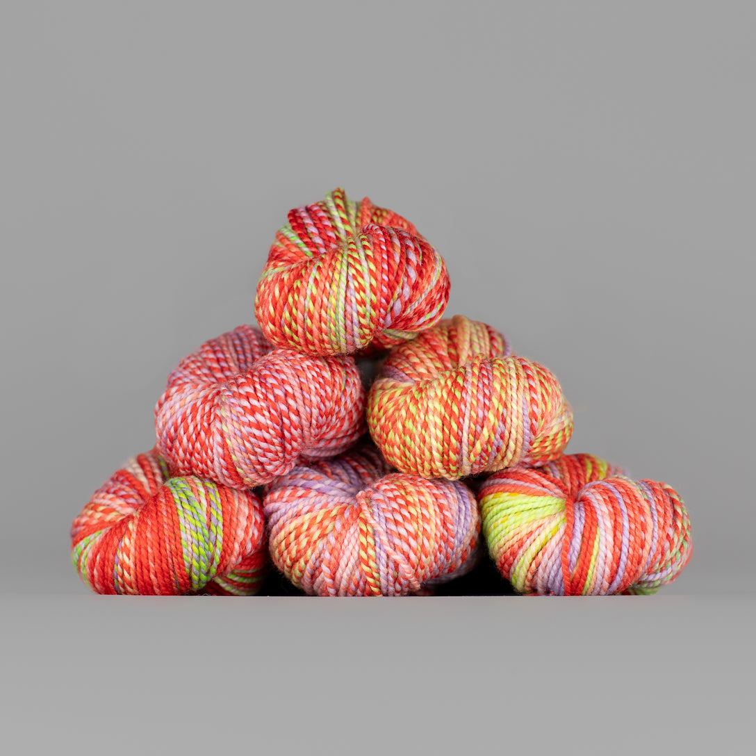 Skeins of Spincycle Dyed in the Wool sport weight yarn in a red orange, pink, and yellow green color gradient against a gray background.
