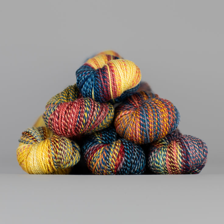 Skeins of Spincycle Dyed in the Wool sport weight yarn in a blue, red, orange, and yellow color gradient against a gray background.