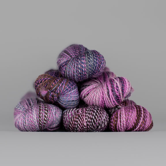 Skeins of Spincycle Dyed in the Wool sport weight yarn in a light and dark purple, brown, and gray color gradient against a gray background.
