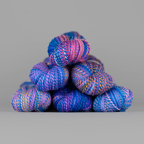 Skeins of Spincycle Dyed in the Wool sport weight yarn in an electric blue, purple, pink, and orange color gradient against a gray background.