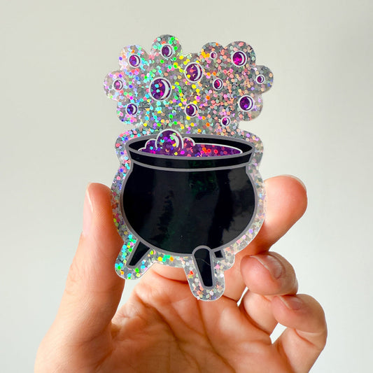 Hand holding a colorful, glittery cauldron sticker a white background