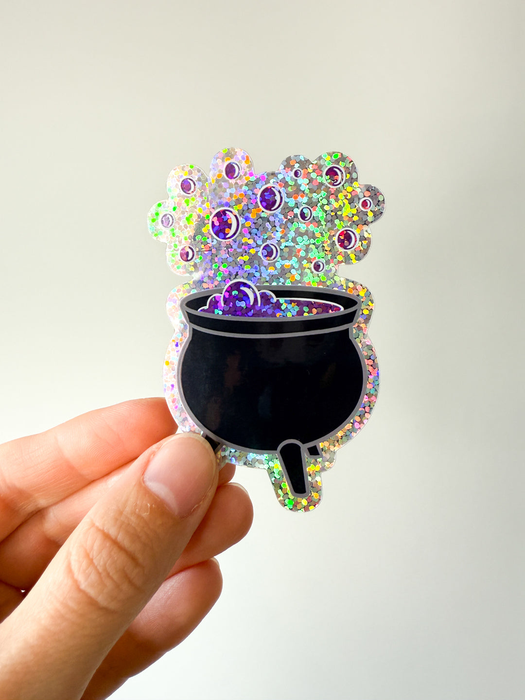 Hand holding a colorful glittery sticker of a cauldron with purple bubbles on a white background