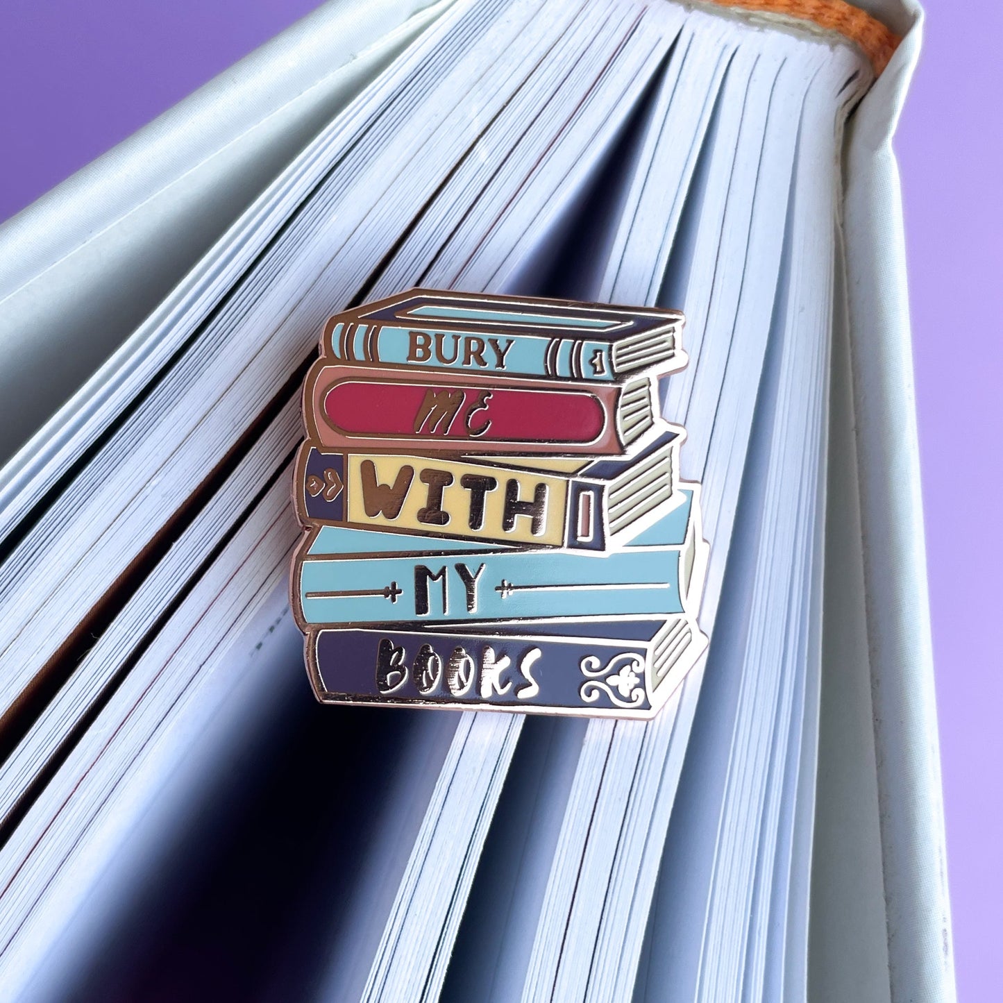 An enamel pin shaped like a stack of books that reads "Bury Me With My Books" on the spine of a book. 