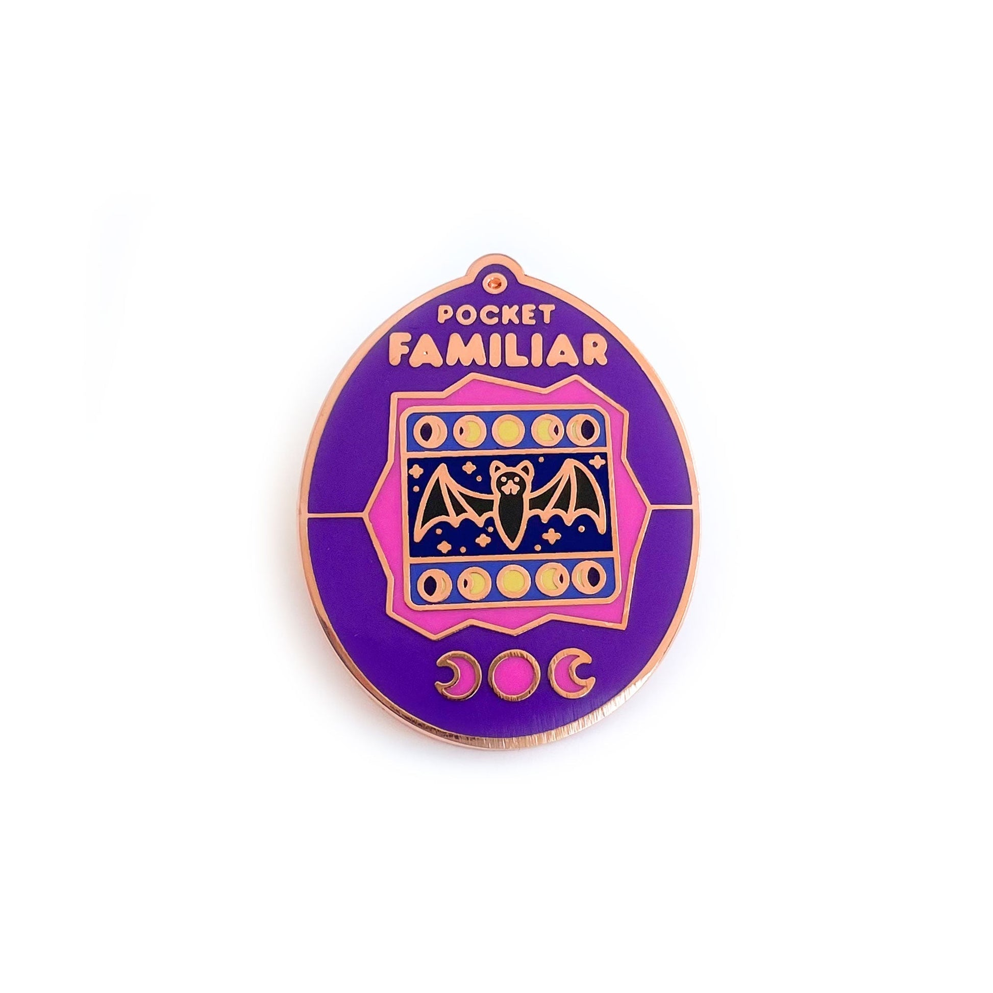 An enamel pin shaped like a Tamagotchi that reads "Pocket Familiar" at the top. It has a pink and purple shell. The screen has moon phases and a bat with night stars by it. The buttons of the tamagotchi are moon phases. 