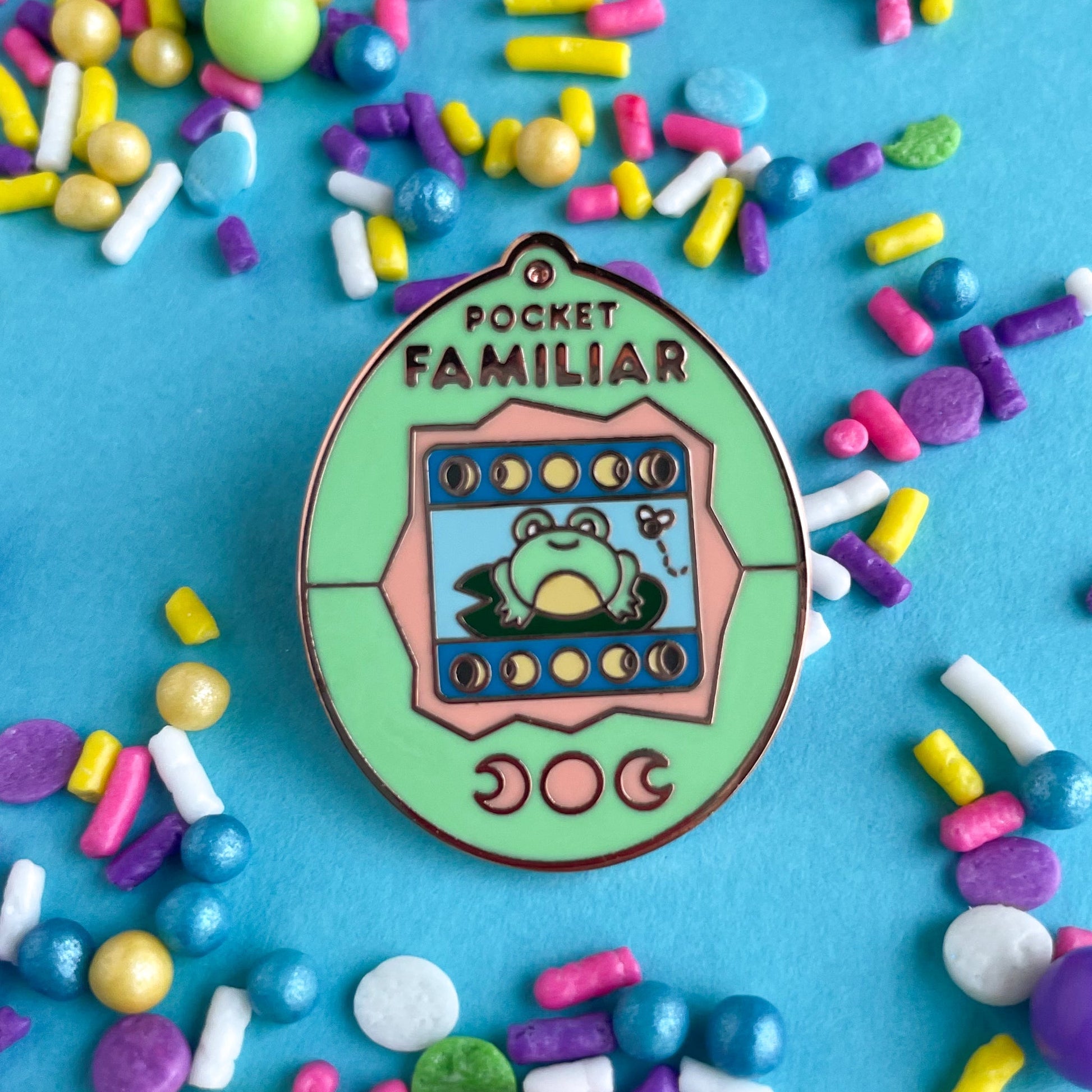 An enamel pin shaped like a Tamagotchi that reads "Pocket Familiar" at the top. It has a mint green and peach shell. The screen has moon phases and a frog on a lily pad looking at a fly. The buttons of the tamagotchi are moon phases. The pin is on a blue background covered in pastel sprinkles.
