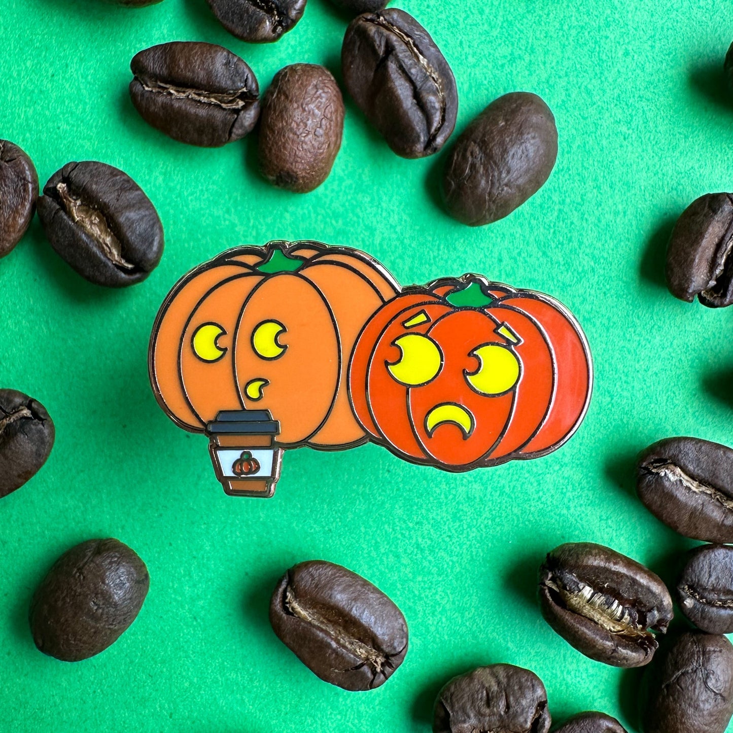 an enamel pin of two pumpkins, one drinking a pumpkin spice latte, the pin is on a green background with coffee beans