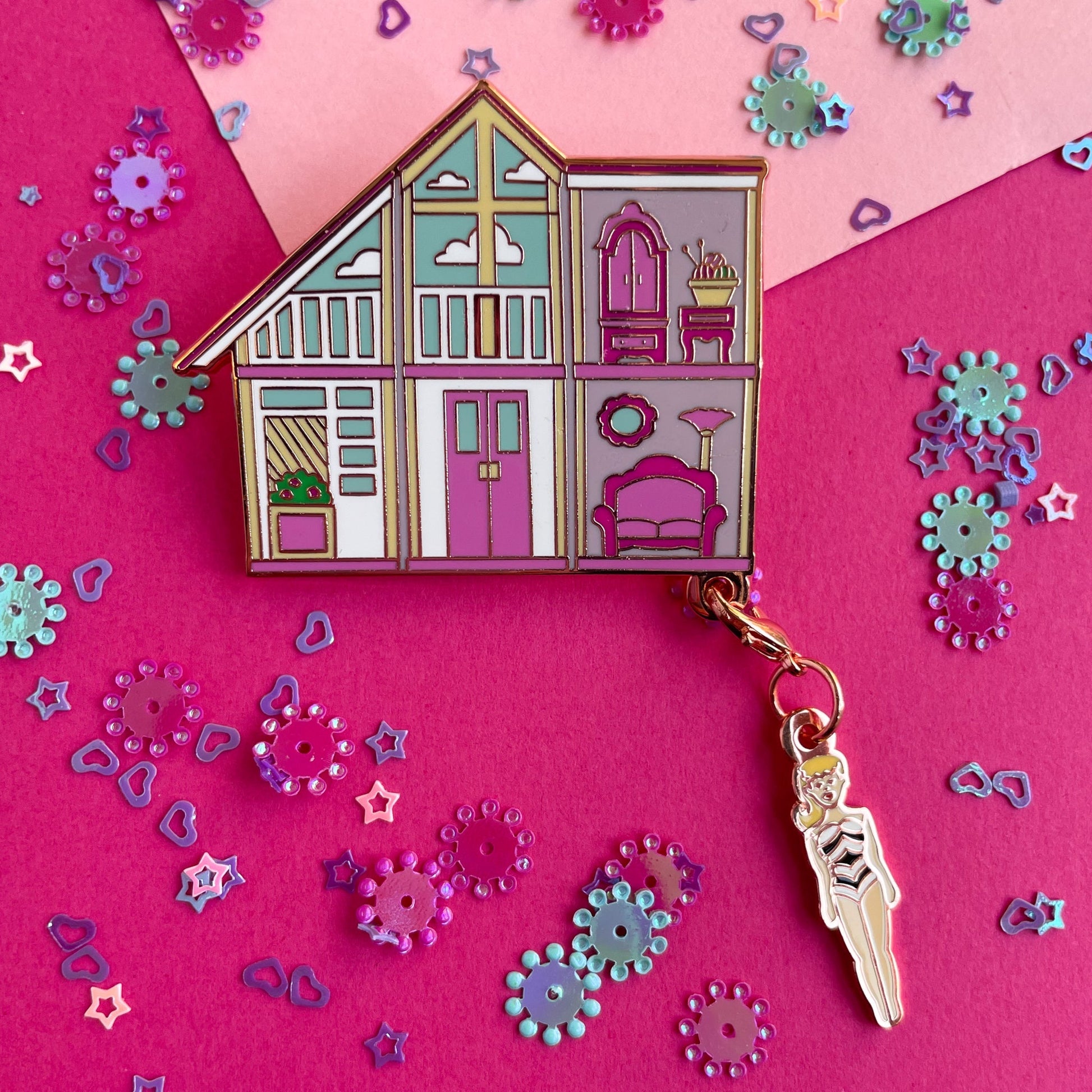 An enamel pin of a pink doll house with a Barbie shaped charm hanging from the corner on a pink paper background with confetti. 