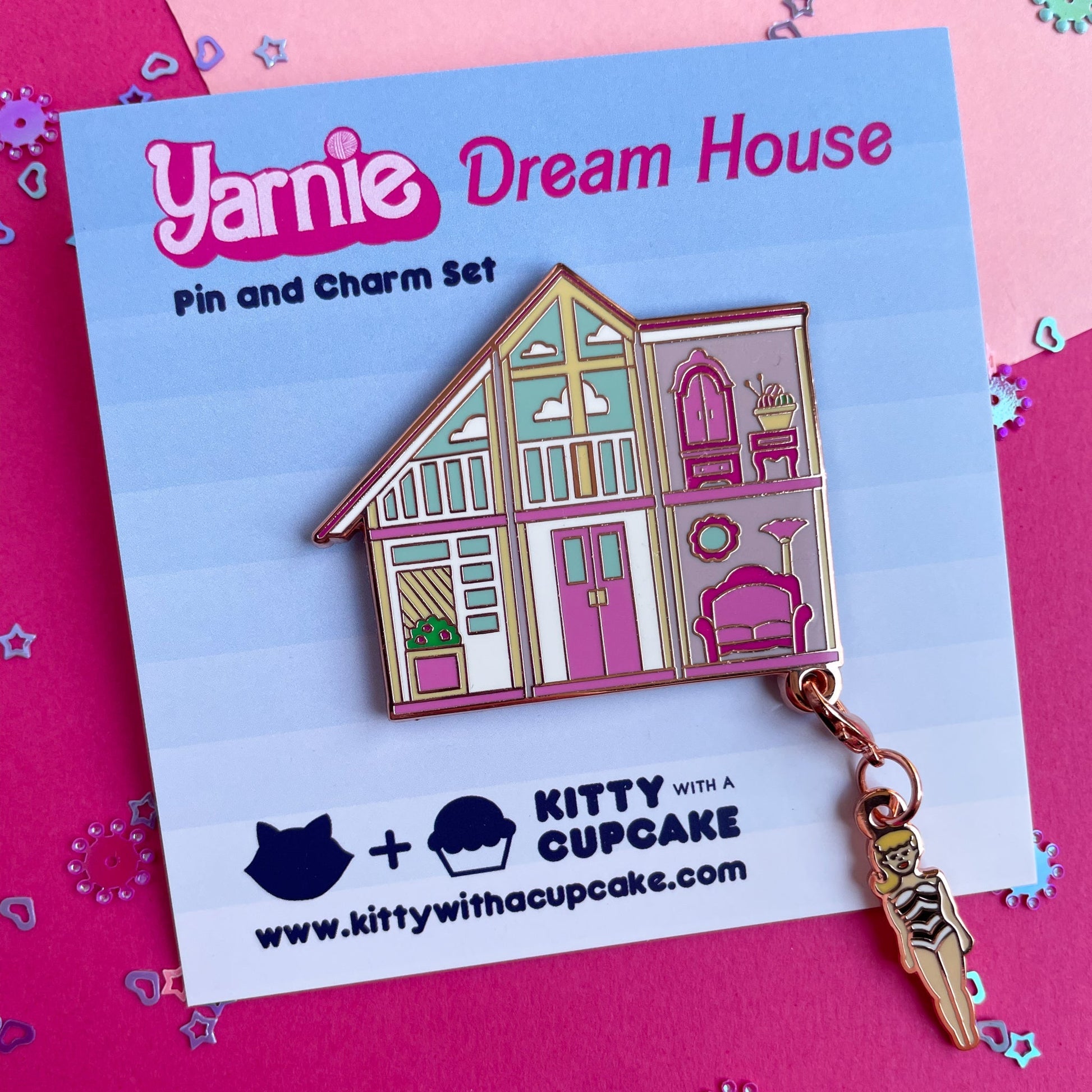 An enamel pin of a pink doll house with a Barbie shaped charm hanging from the corner on a blue backing card that reads "Yarnie Dream House"