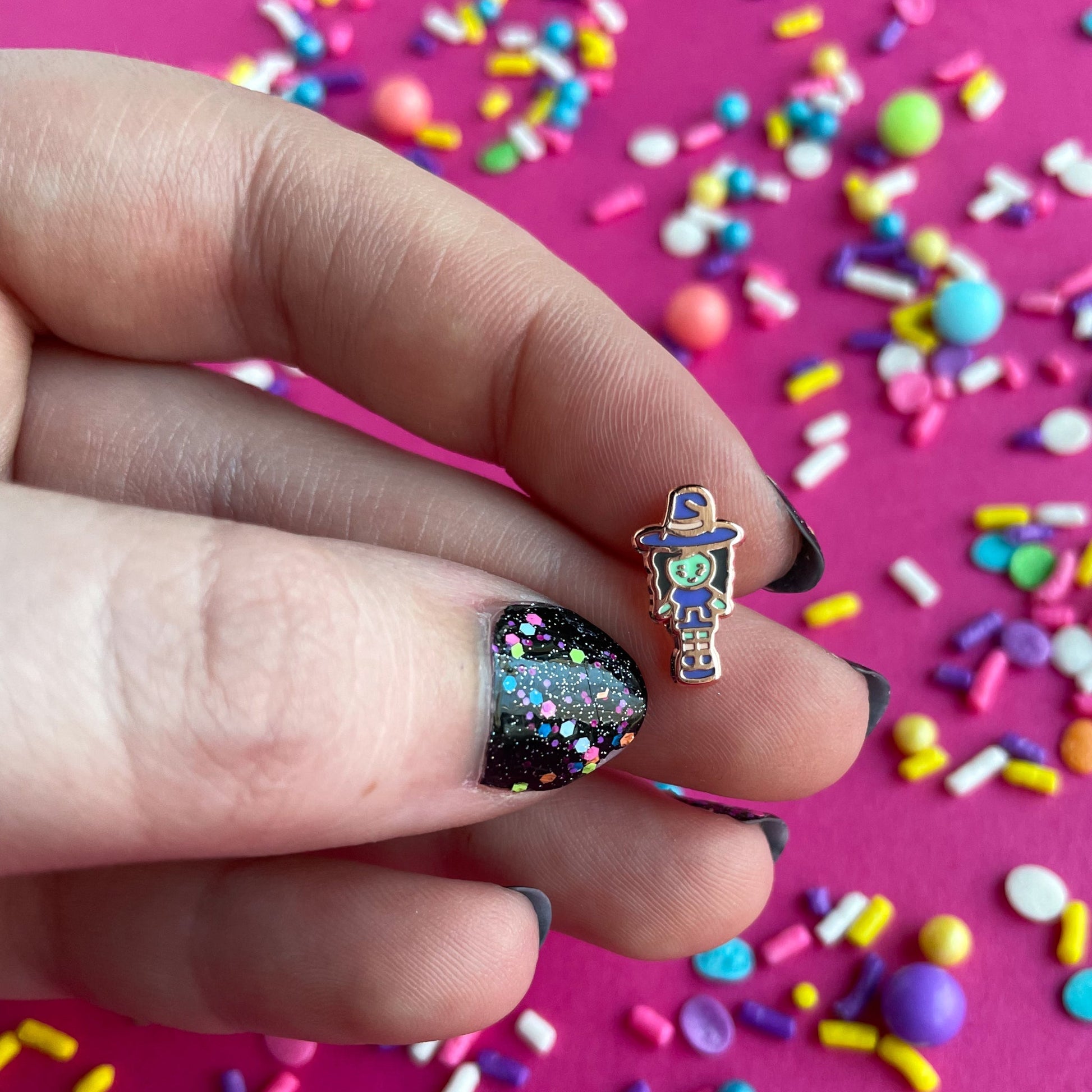 A close up of a hand with black and confetti painted nails holding the Witchy Pocket pin. She is a small polly pocket style doll about the size of a fingernail. She is wearing a purple dress and hat. She has green skin.