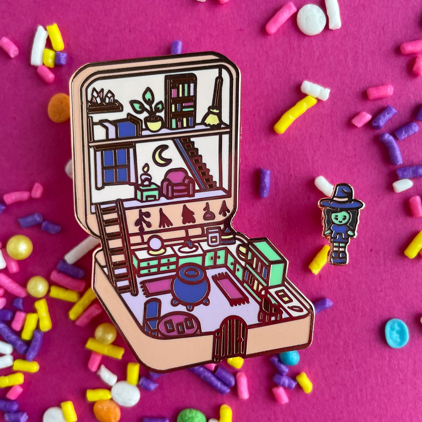 A pin set that is a Polly Pocket style compact of a witch house with a little witch mini doll. The compact house has a cauldron, crystal ball, tarot cards, crystals, and a broom. The compact is a pastel peach color with mint green, hot pink and purple details. The pins are on a hot pink background that is covered in pastel sprinkles.