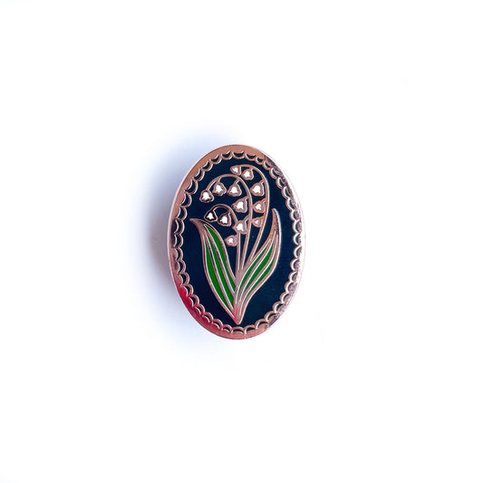 An oval shaped enamel pin with a lily of the valley flower on it. 