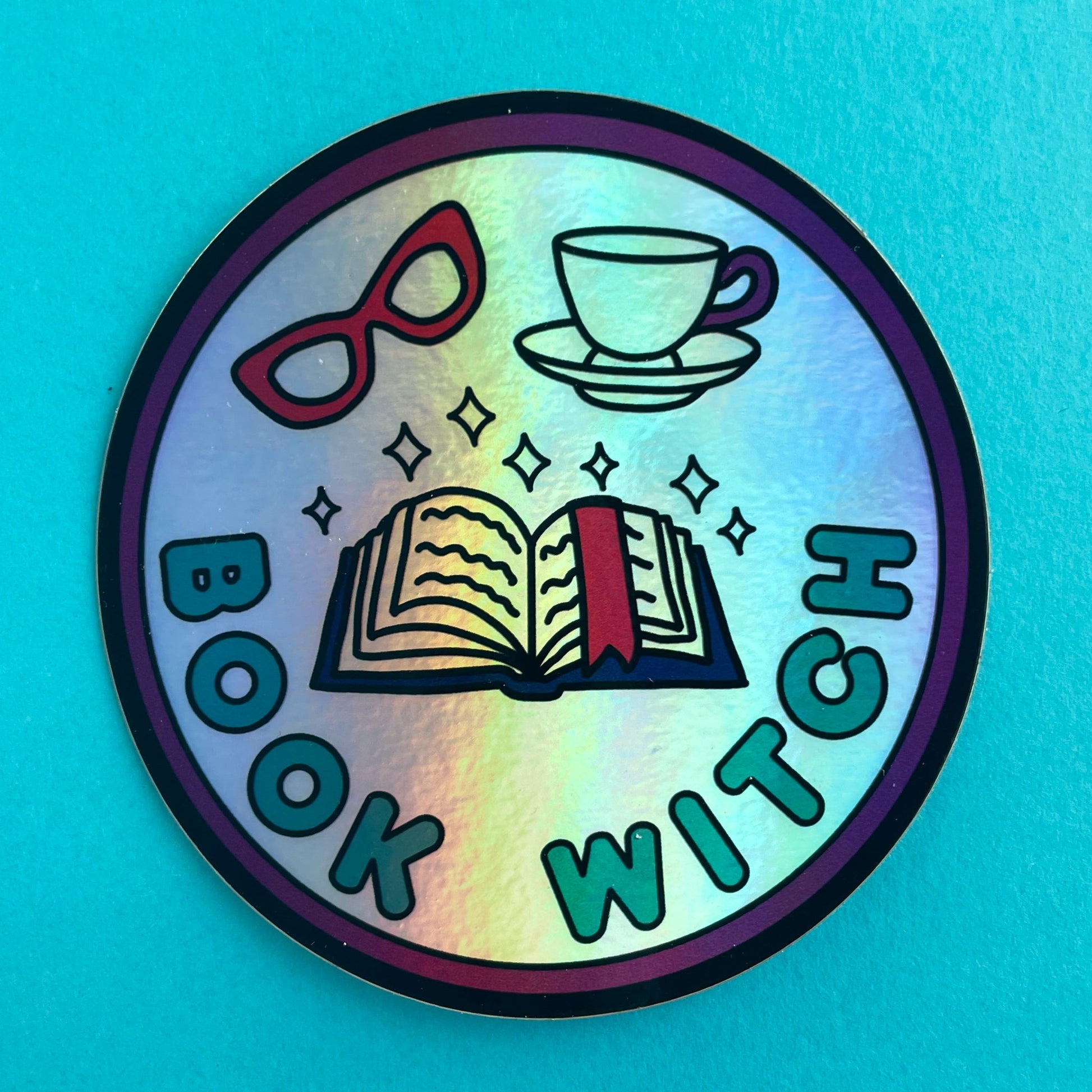 A circular holographic sticker. It has a maroon border with turquoise text that reads "Book Witch". There is an open book, sparkles, red glasses and a teacup above the words.