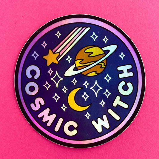 A circular holographic sticker. It has a hot pink border and a dark blue background with light pink words that read "Cosmic Witch". Depicted above the words are a crescent moon, sparkle stars, a shooting star, and the planet Saturn. The sticker is on a hot pink background.