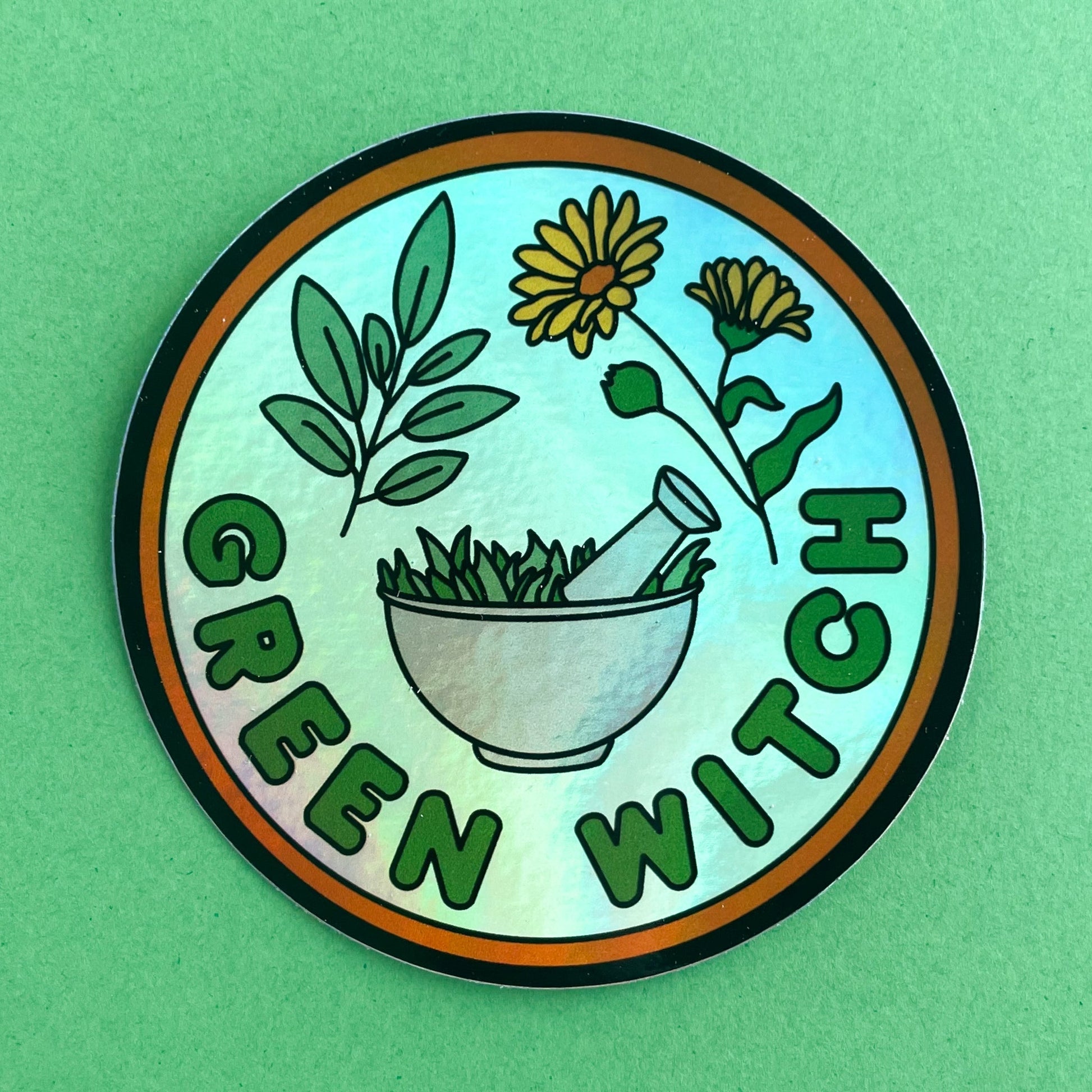 A circular holographic sticker that has an orange border and the words "Green Witch" on it. Sage leaves, calendula flowers, and a mortar and pestle are depicted on it. The sticker is on a green background.