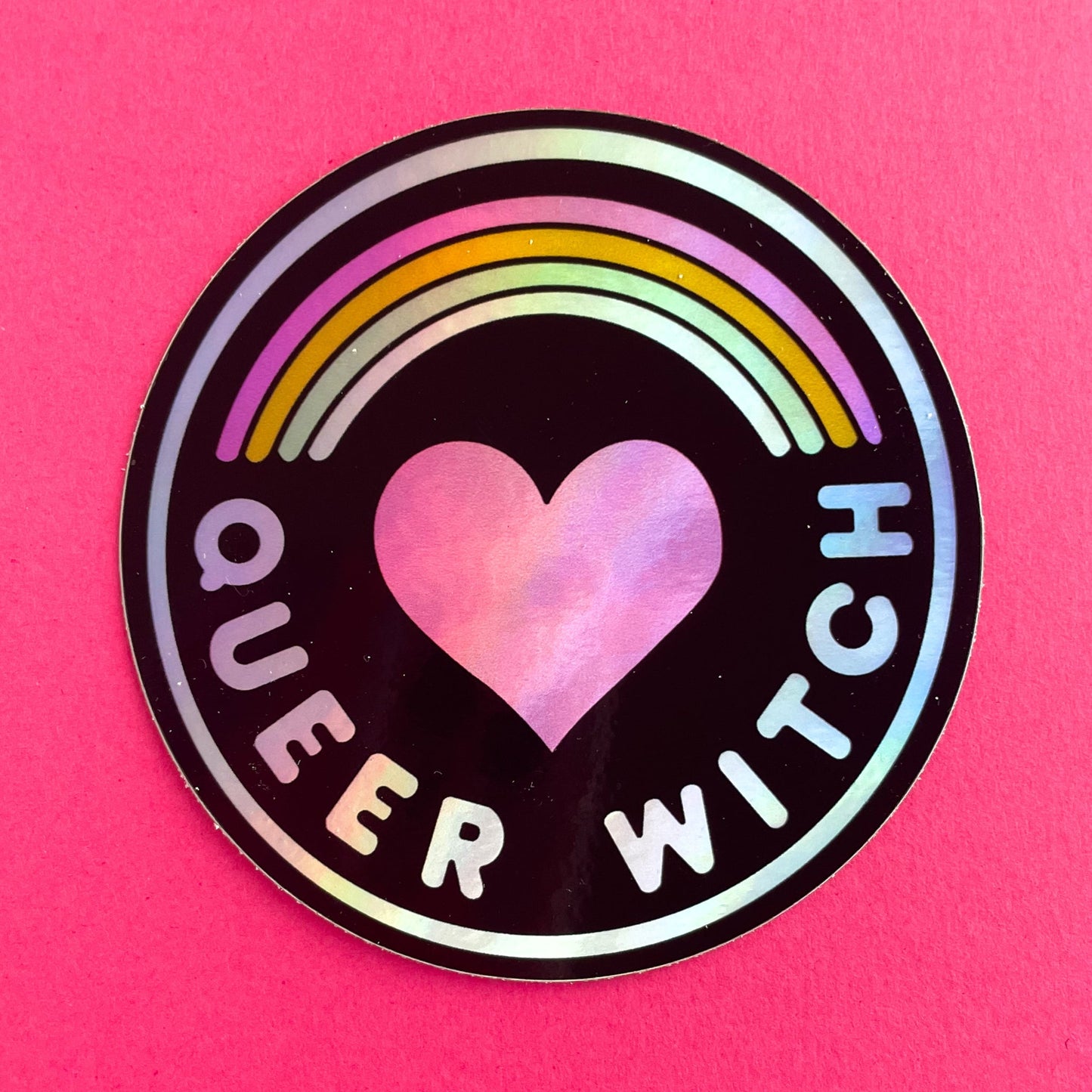 A circular holographic sticker with a black background and the words "Queer Witch" on it. It has a heart with a rainbow above it. The sticker is on a hot pink background.