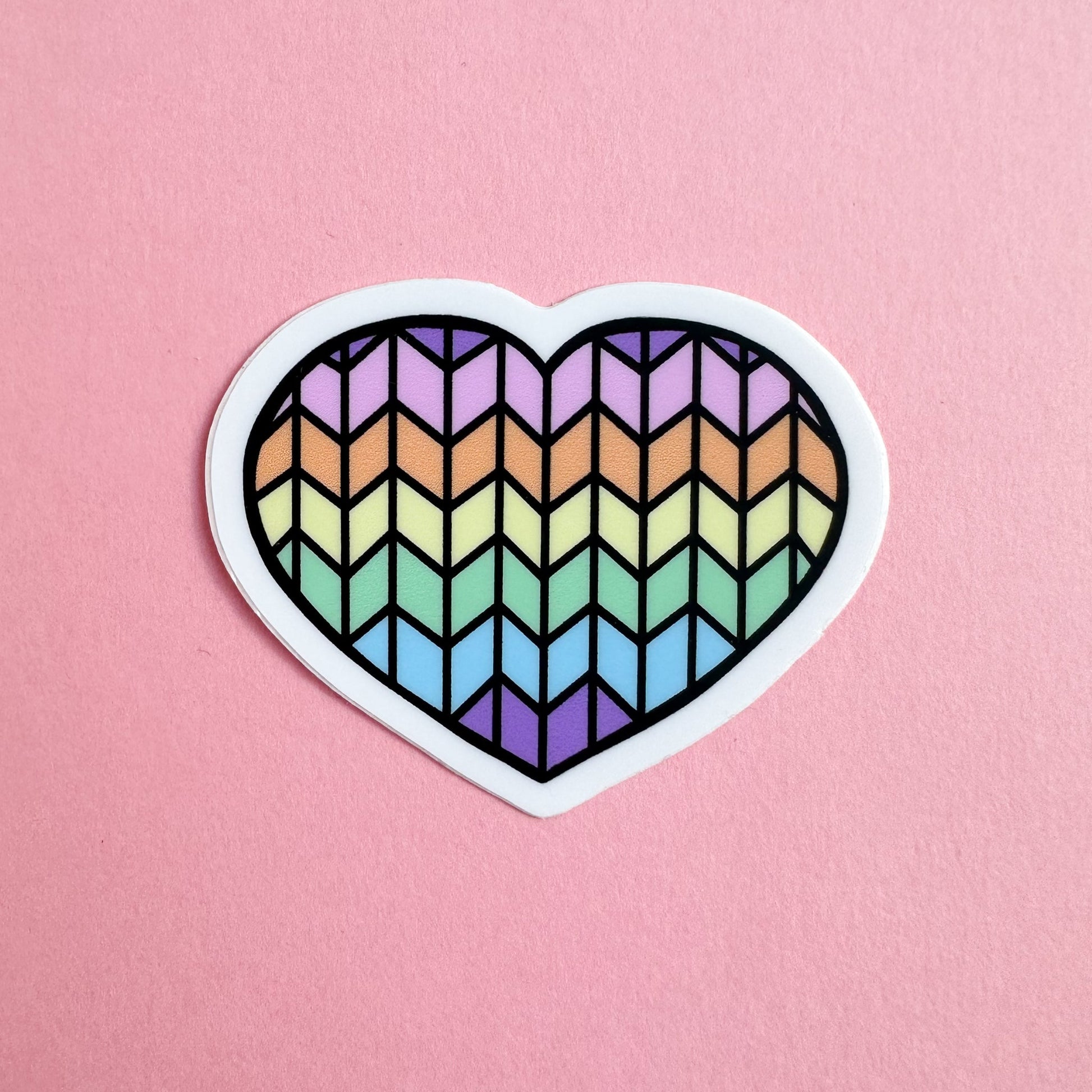 A heart shaped vinyl sticker with a pastel rainbow stockinette stitch pattern on it on a pink background