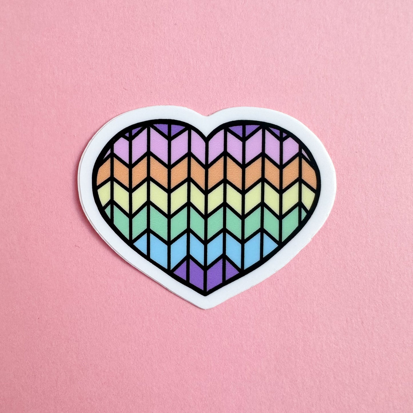 A heart shaped vinyl sticker with a pastel rainbow stockinette stitch pattern on it on a pink background