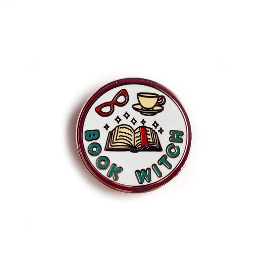 A circular enamel pin in the style of a merit badge featuring illustrations of an open book with sparkles, a teacup, and red glasses. Underneath the illustrations are the words "Book Witch" in teal bubble letters. 