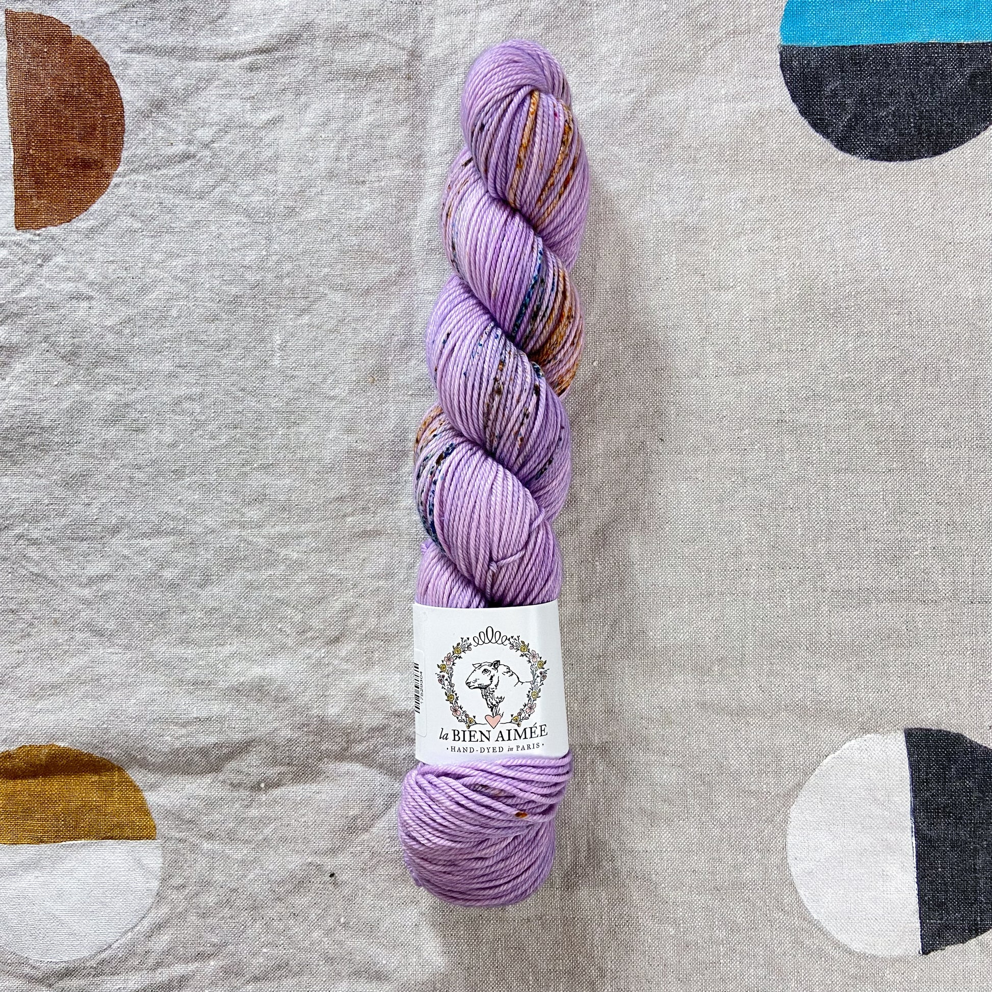 An image of a skein of DK weight yarn in a light purple color with speckles of teal and brown, with a white ‘La Bien Aimee’ label against a canvas background.