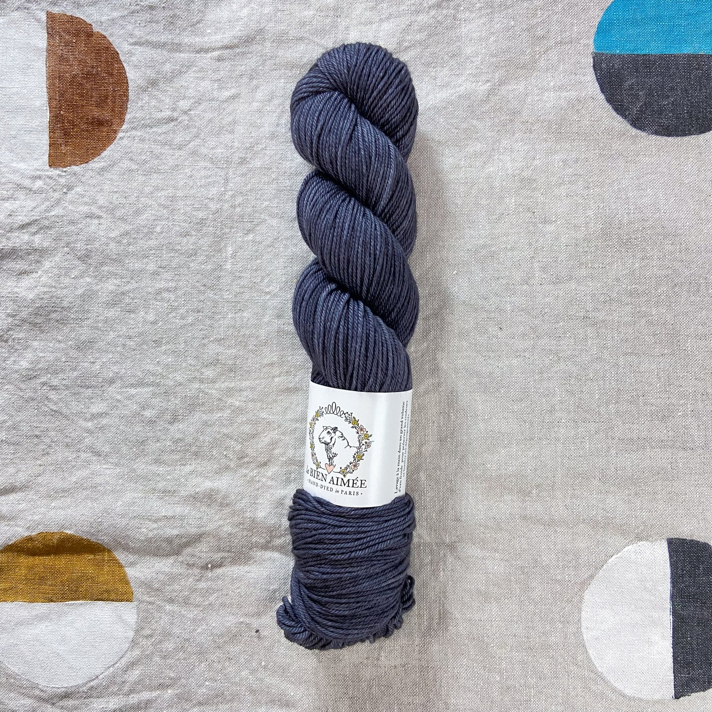 An image of a skein of DK weight yarn in a charcoal gray color with a white ‘La Bien Aimee’ label against a canvas background.