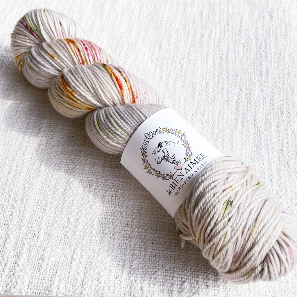 An image of a skein of DK weight yarn in a cream color with speckles of pink, orange, and green, with a white ‘La Bien Aimee’ label against a canvas background.