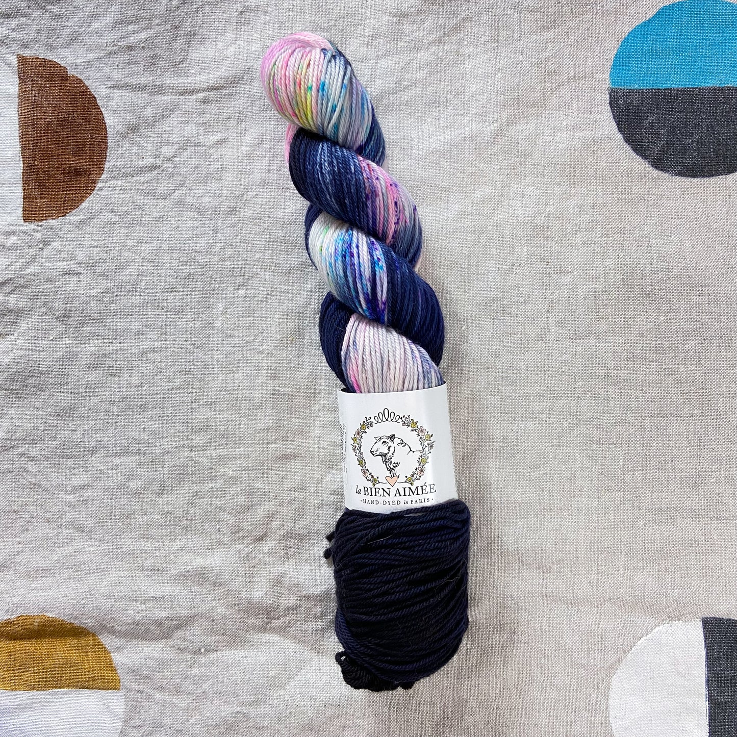 An image of a skein of DK weight yarn in navy blue, pale pink, yellow, and gray colors.with a white ‘La Bien Aimee’ label against a canvas background.