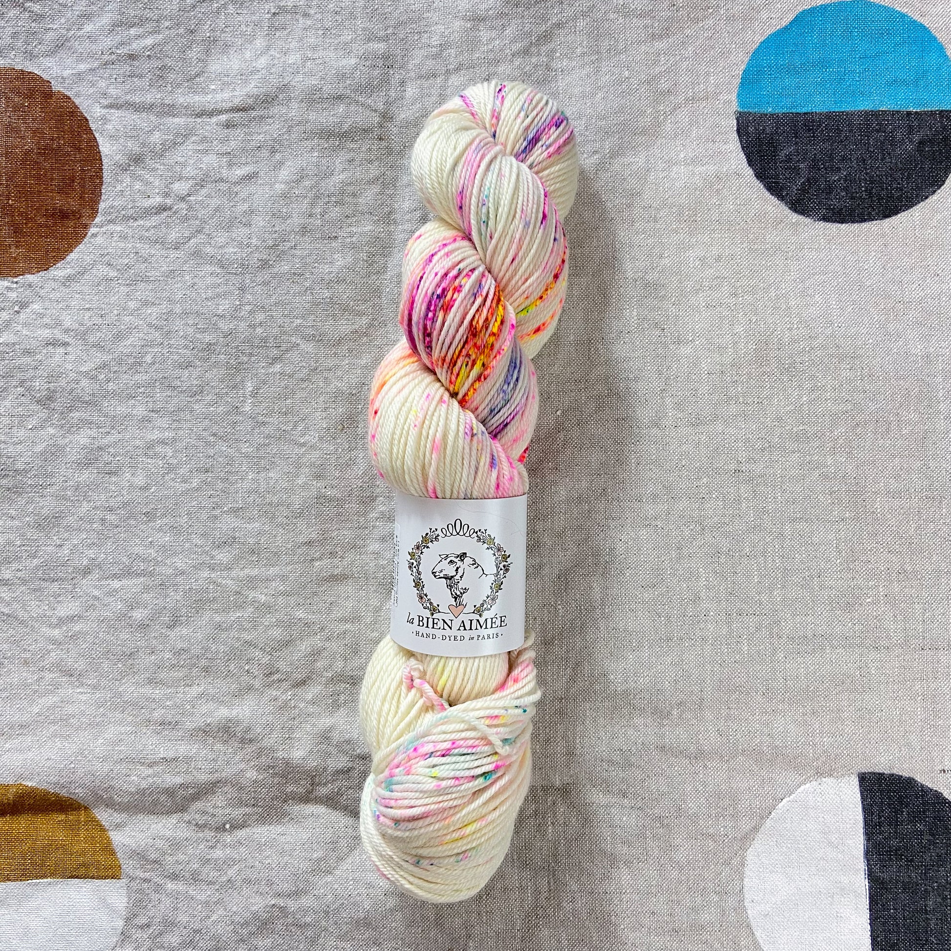 An image of a skein of DK weight yarn in a cream color with speckles of bright pink, orange, blue, purple, and yellow, with a white ‘La Bien Aimee’ label against a canvas background.