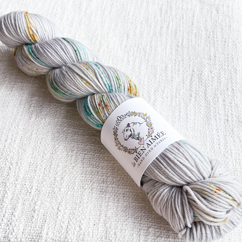 An image of a skein of DK weight yarn in a pale gray color with speckles of teal and yellow, with a white ‘La Bien Aimee’ label against a canvas background.