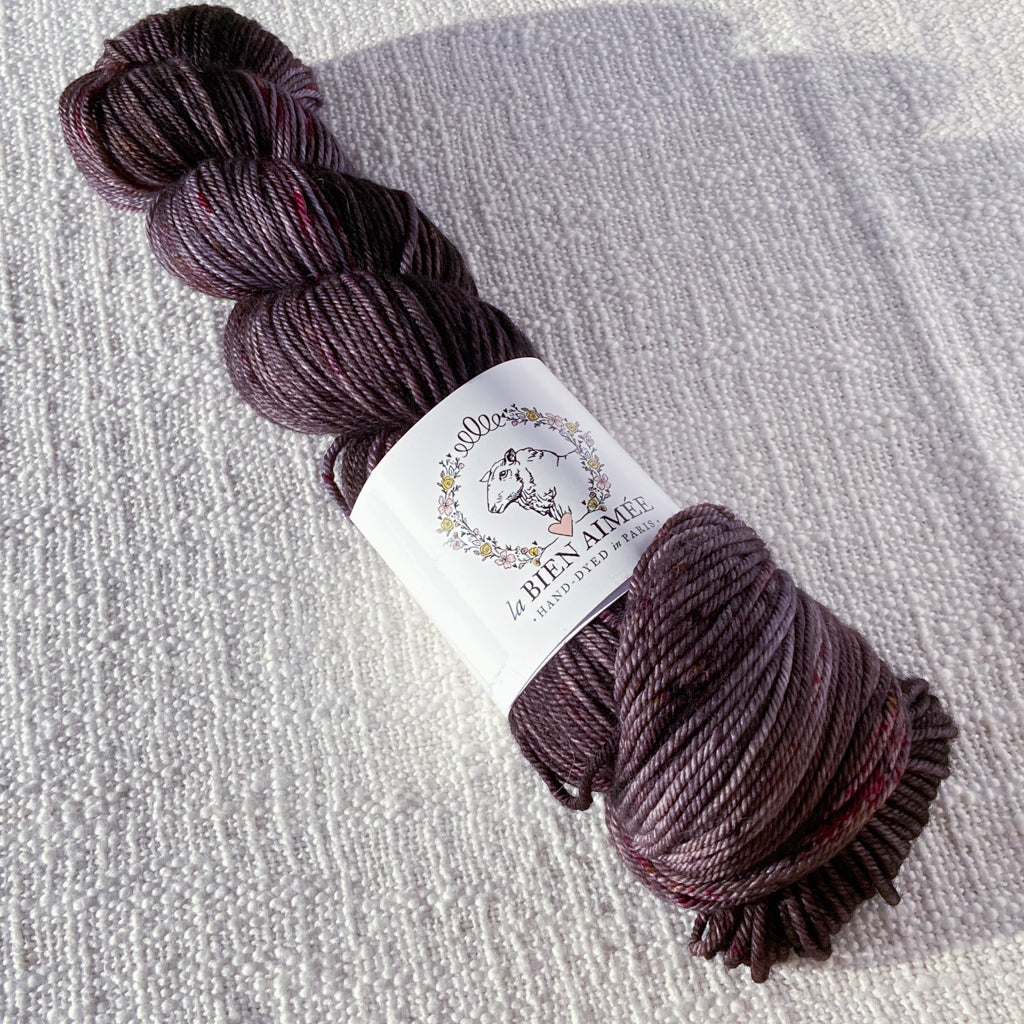 An image of a skein of DK weight yarn in a deep purple brown color with speckles of magenta, with a white ‘La Bien Aimee’ label against a canvas background.