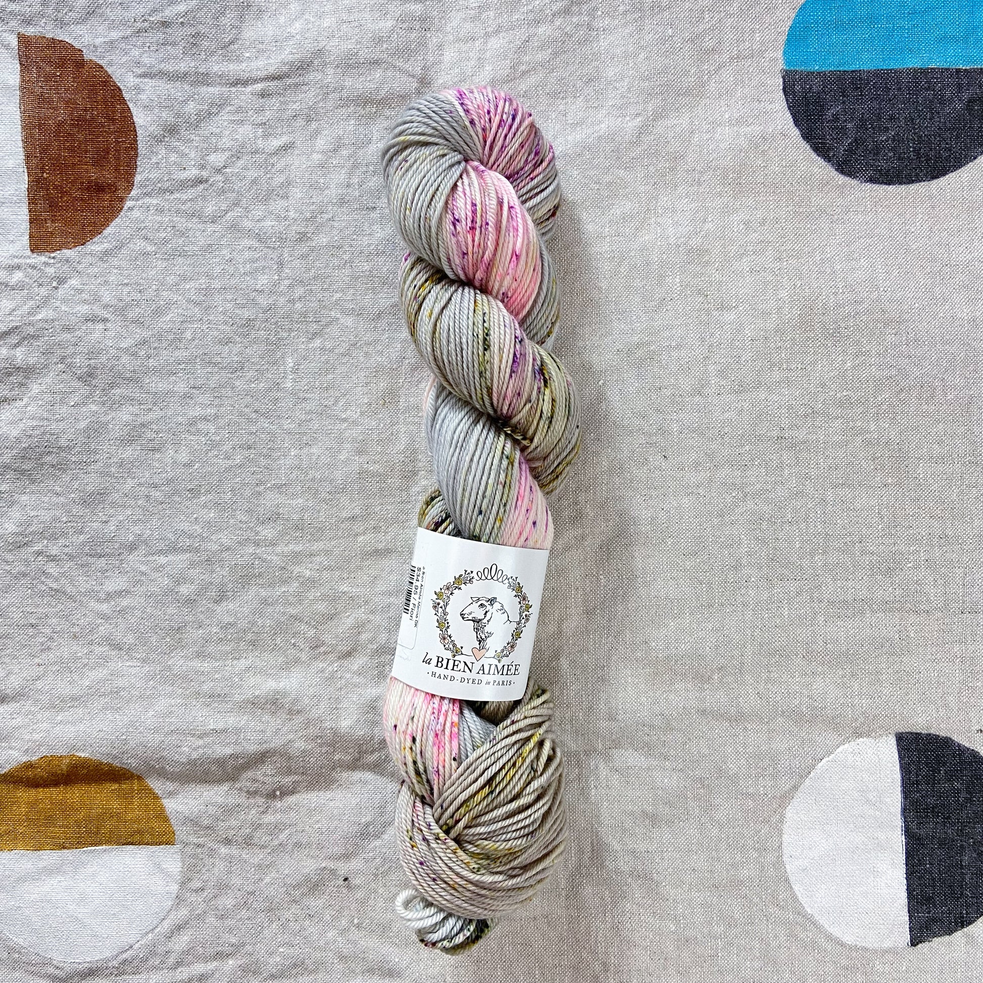 An image of a skein of DK weight yarn in pink, green, and gray colors with a white ‘La Bien Aimee’ label against a canvas background.