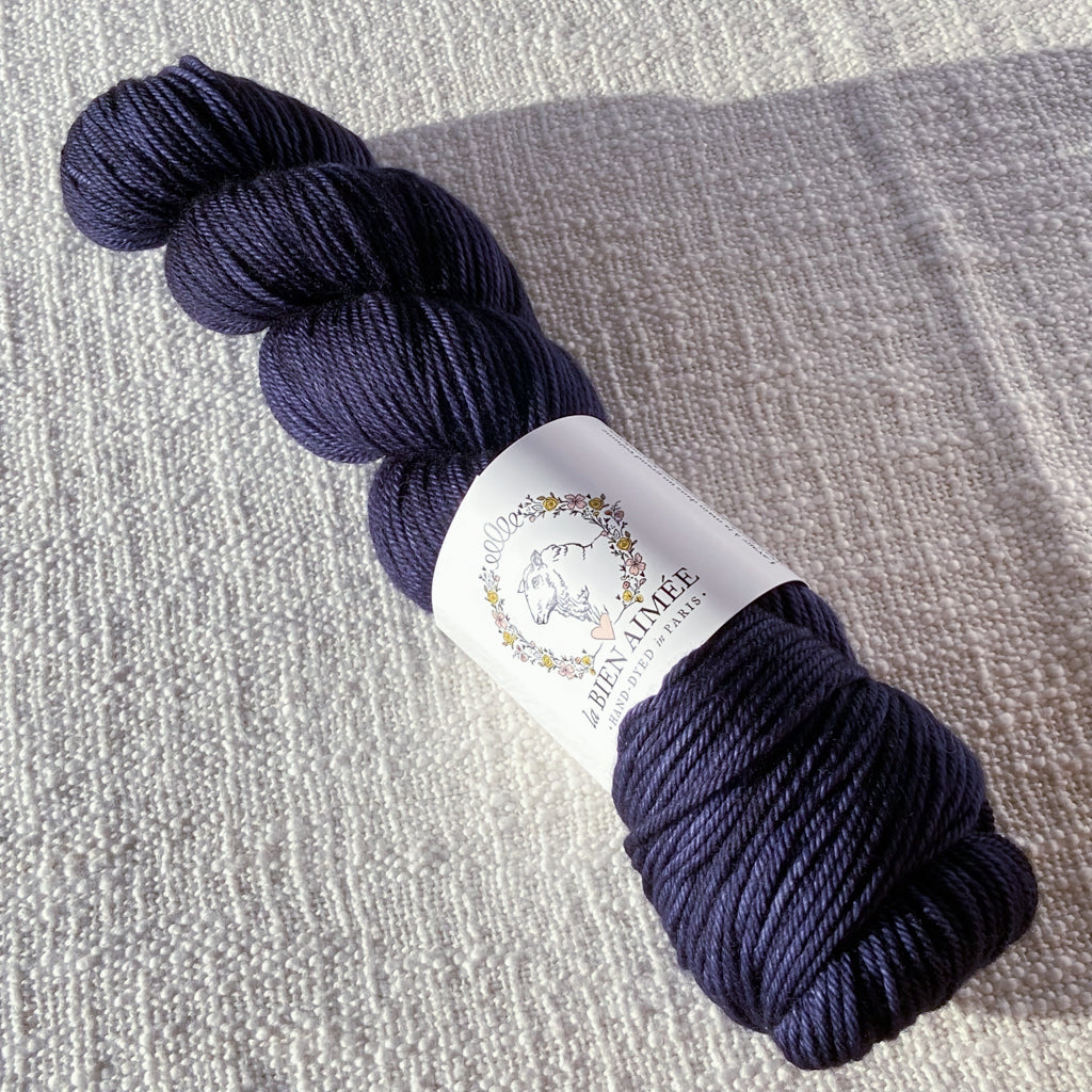 An image of a skein of DK weight yarn in a deep indigo color with a white ‘La Bien Aimee’ label against a canvas background.