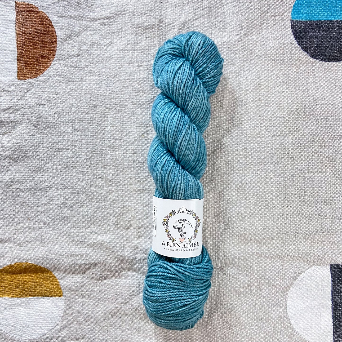 An image of a skein of DK weight yarn in a bright blue color with a white ‘La Bien Aimee’ label against a canvas background.