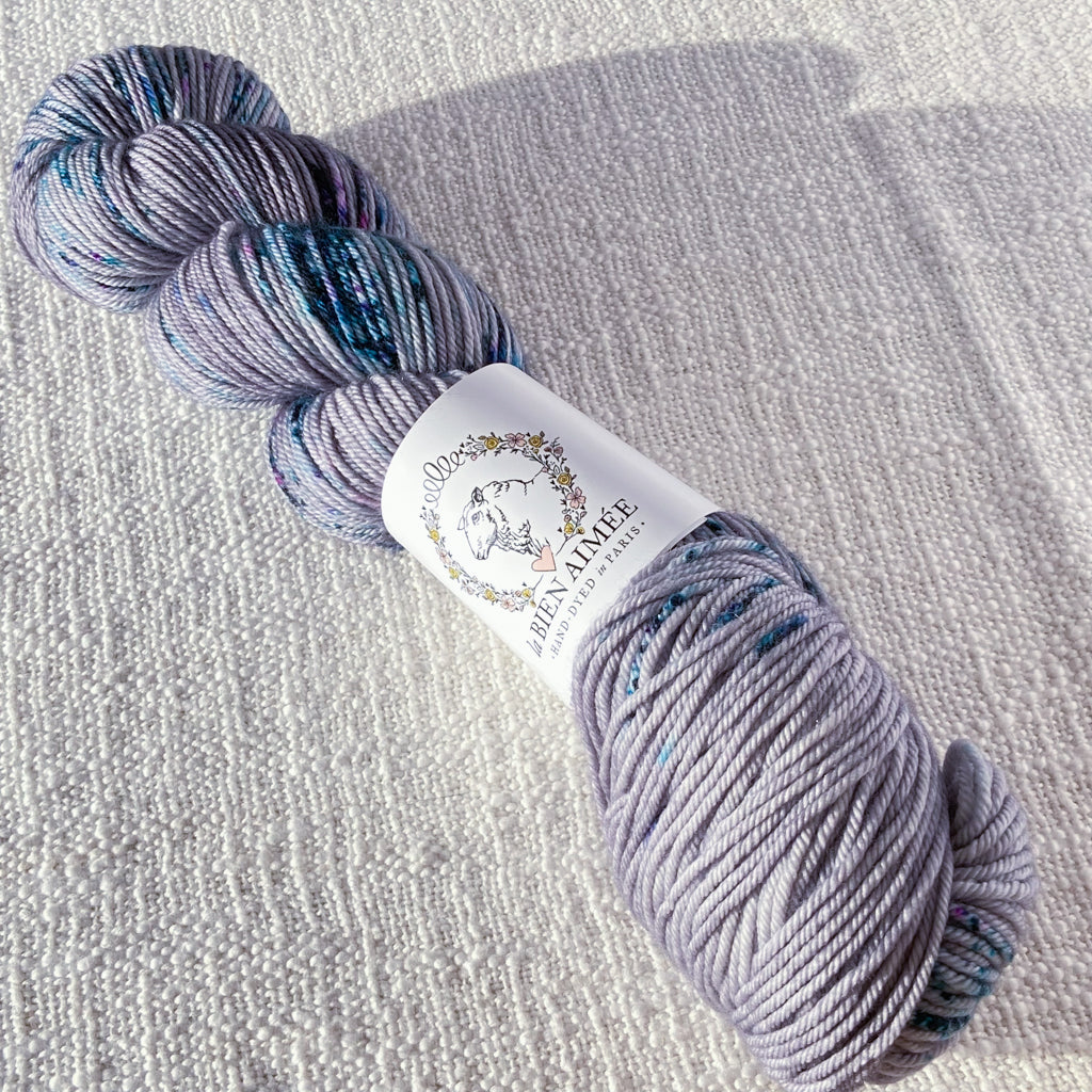 An image of a skein of DK weight yarn in a light purple gray color with speckles of blue and purple, with a white ‘La Bien Aimee’ label against a canvas background.