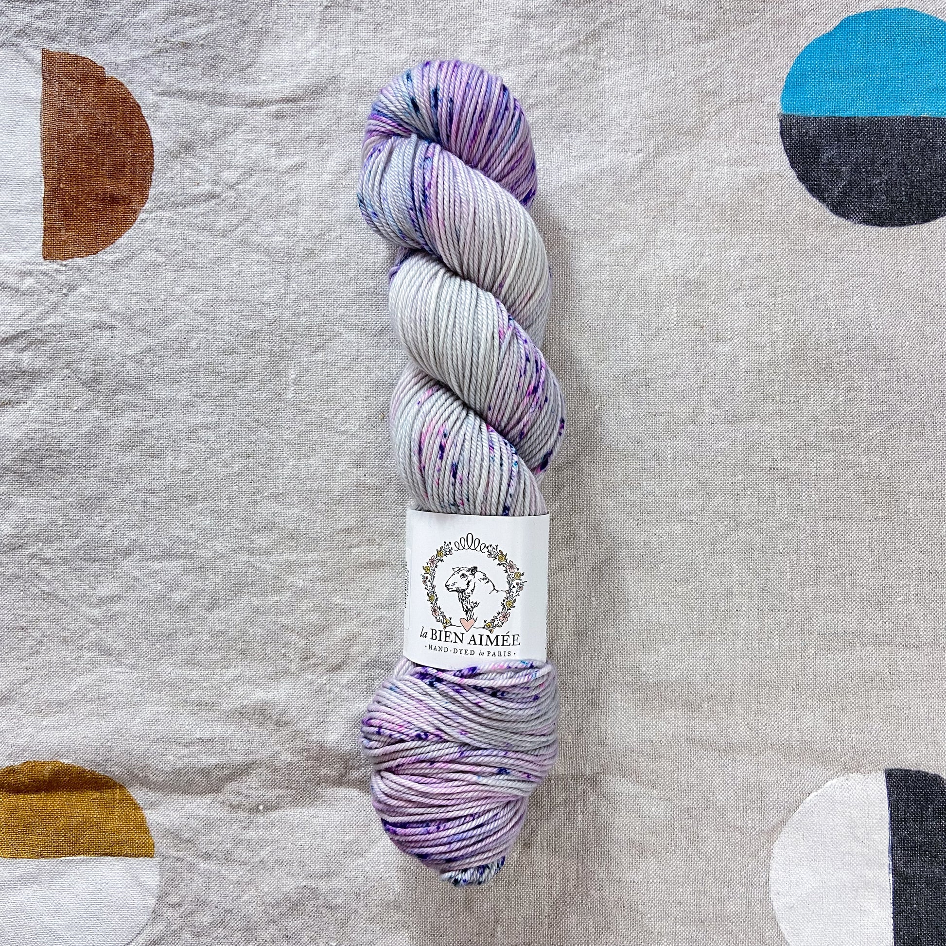 An image of a skein of DK weight yarn in shades of light purple and gray with speckles of blue and purple, with a white ‘La Bien Aimee’ label against a canvas background.