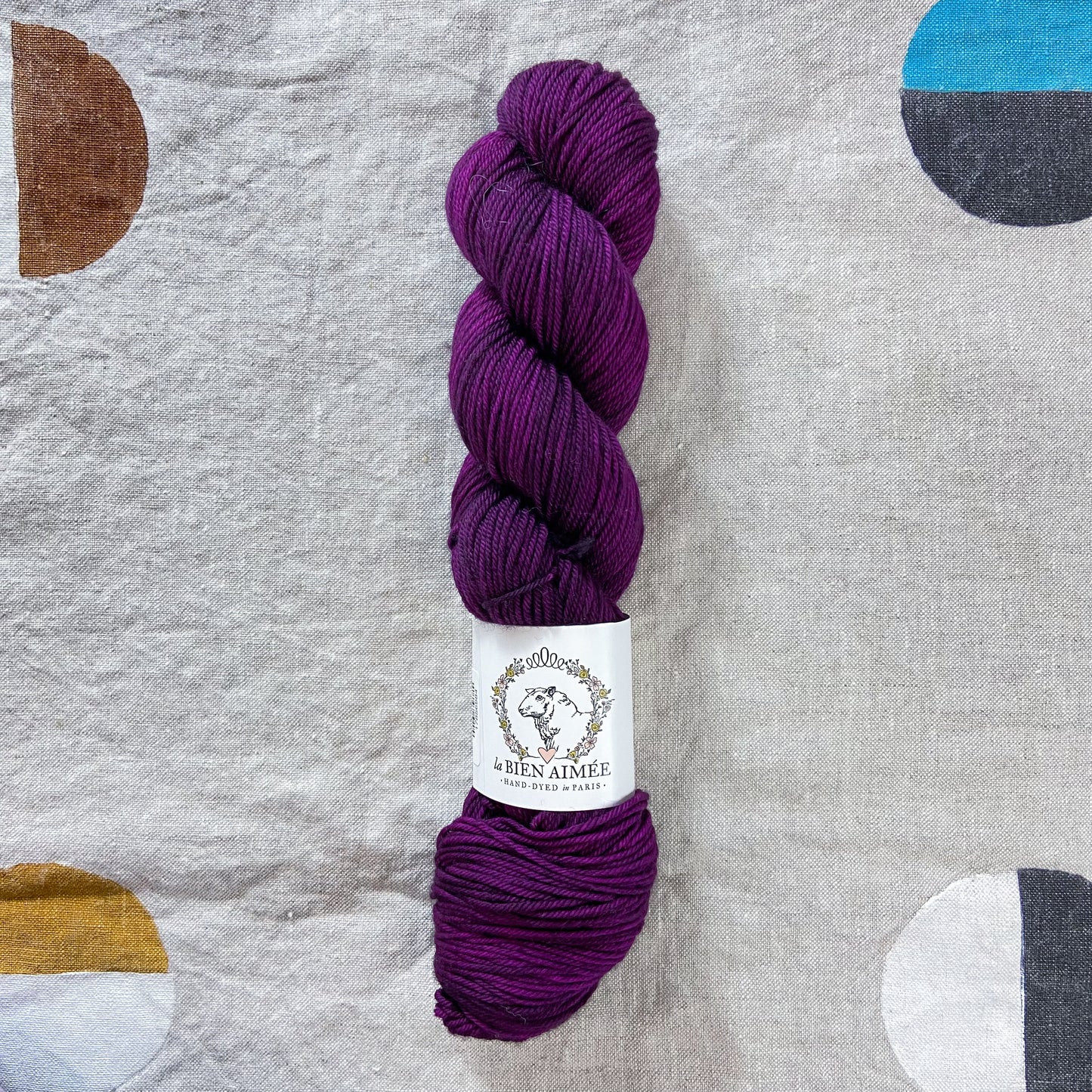 An image of a skein of DK weight yarn in shades of dark purple with a white ‘La Bien Aimee’ label against a canvas background.