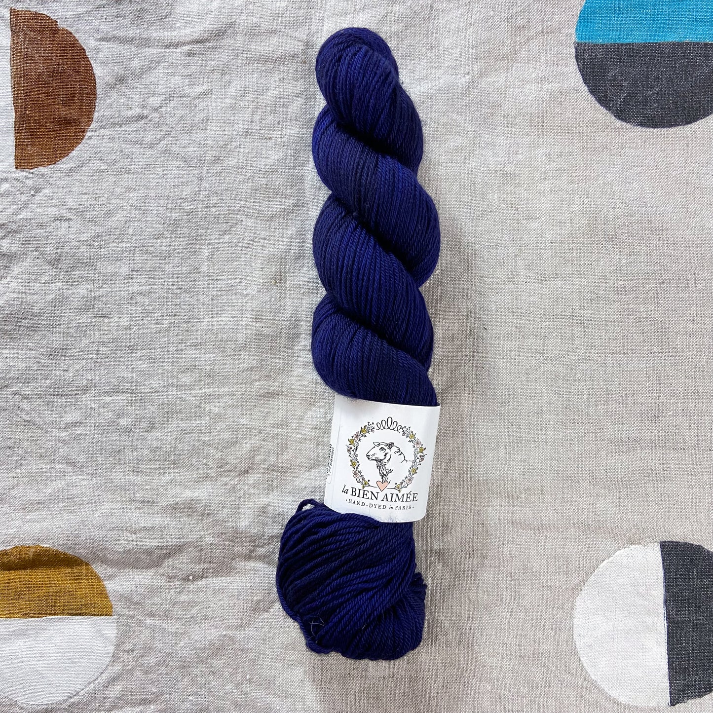 An image of a skein of DK weight yarn in a deep navy blue color with a white ‘La Bien Aimee’ label against a canvas background.