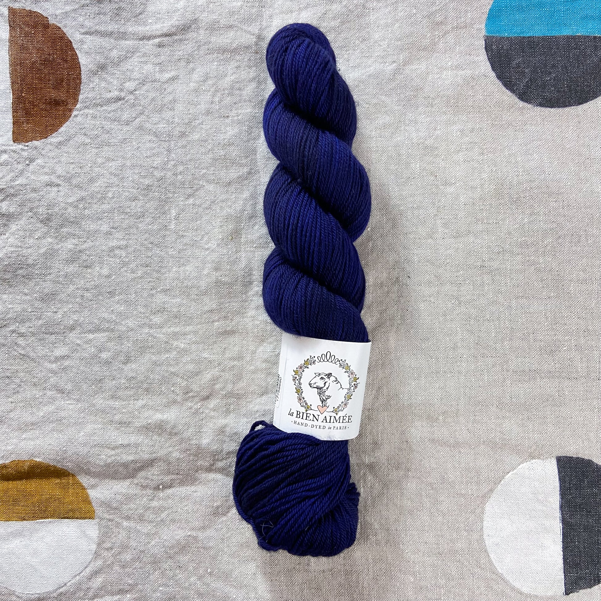 An image of a skein of DK weight yarn in a deep navy blue color with a white ‘La Bien Aimee’ label against a canvas background.