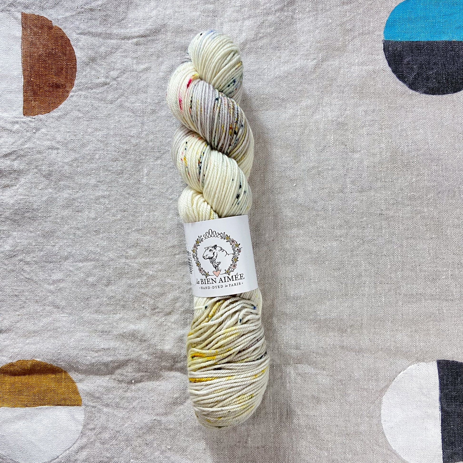 An image of a skein of DK weight yarn in cream, gray, and yellow shades with teal and pink speckles, with a white ‘La Bien Aimee’ label against a canvas background.