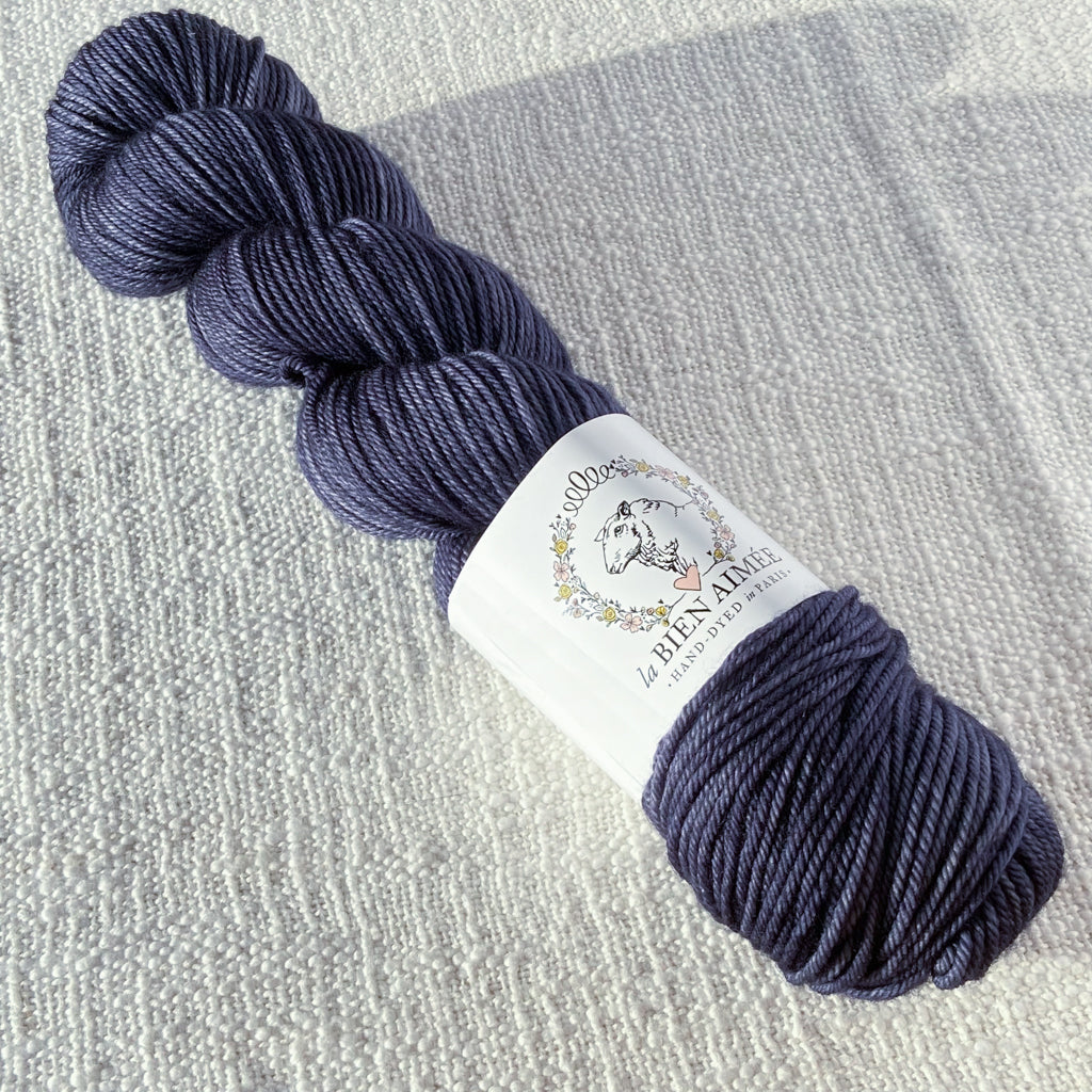 An image of a skein of DK weight yarn in a slate grey color with a white ‘La Bien Aimee’ label against a canvas background.