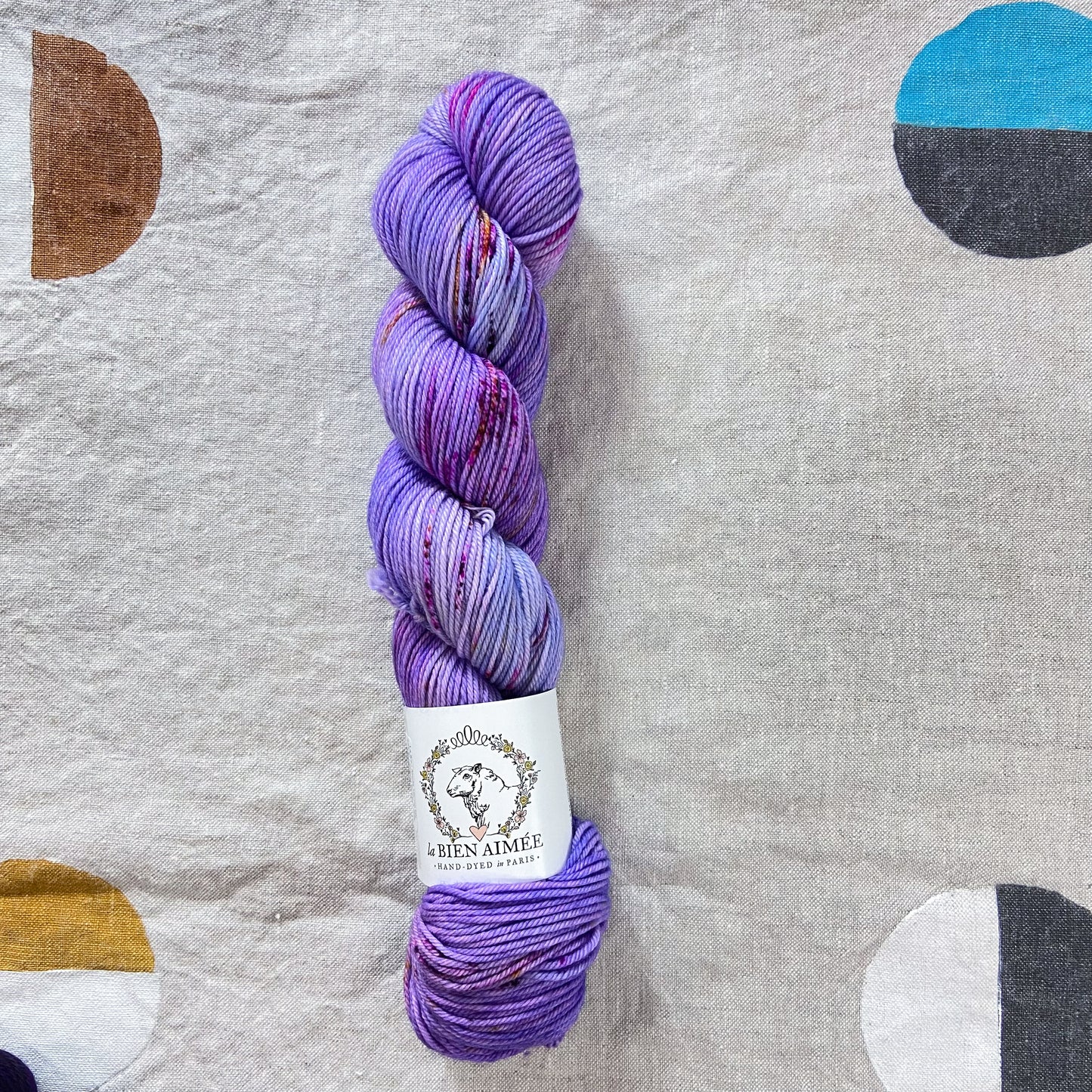 An image of a skein of DK weight yarn in shades of purple with pink and brown speckles, with a white ‘La Bien Aimee’ label against a canvas background.