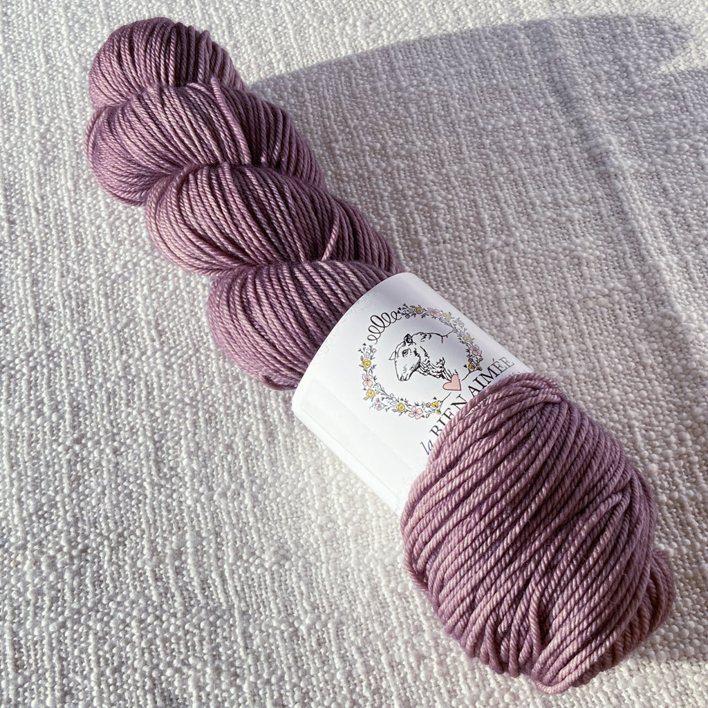 An image of a skein of DK weight yarn in a mauve color with a white ‘La Bien Aimee’ label against a canvas background.