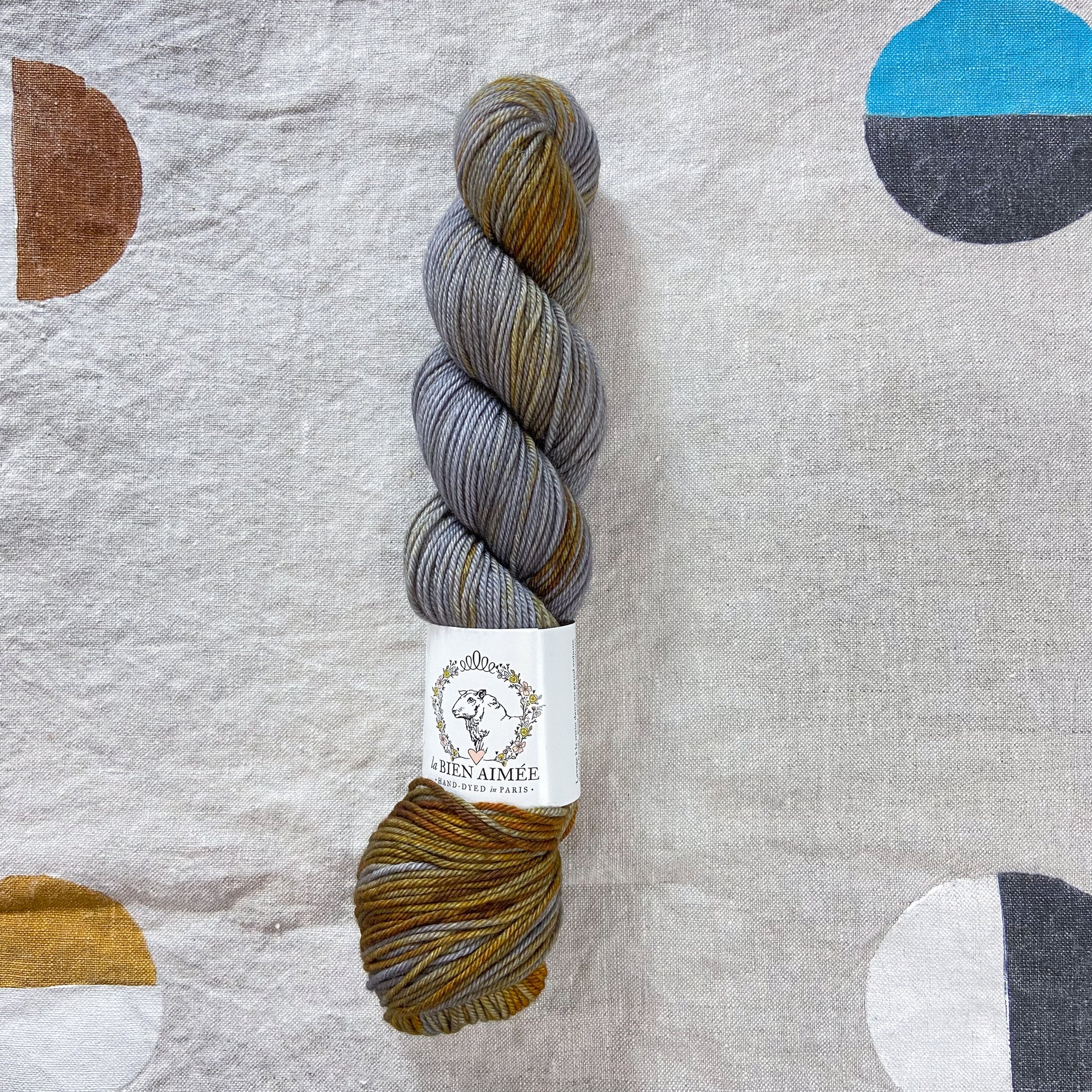 An image of a skein of DK weight yarn in shades of gray and orange with a white ‘La Bien Aimee’ label against a canvas background.