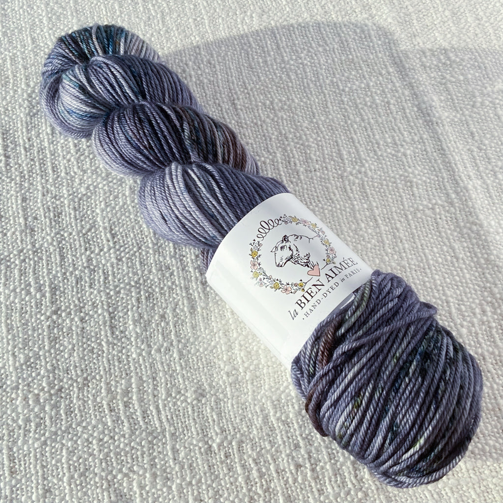 An image of a skein of DK weight yarn in shades of purple gray and blue with a white ‘La Bien Aimee’ label against a canvas background.