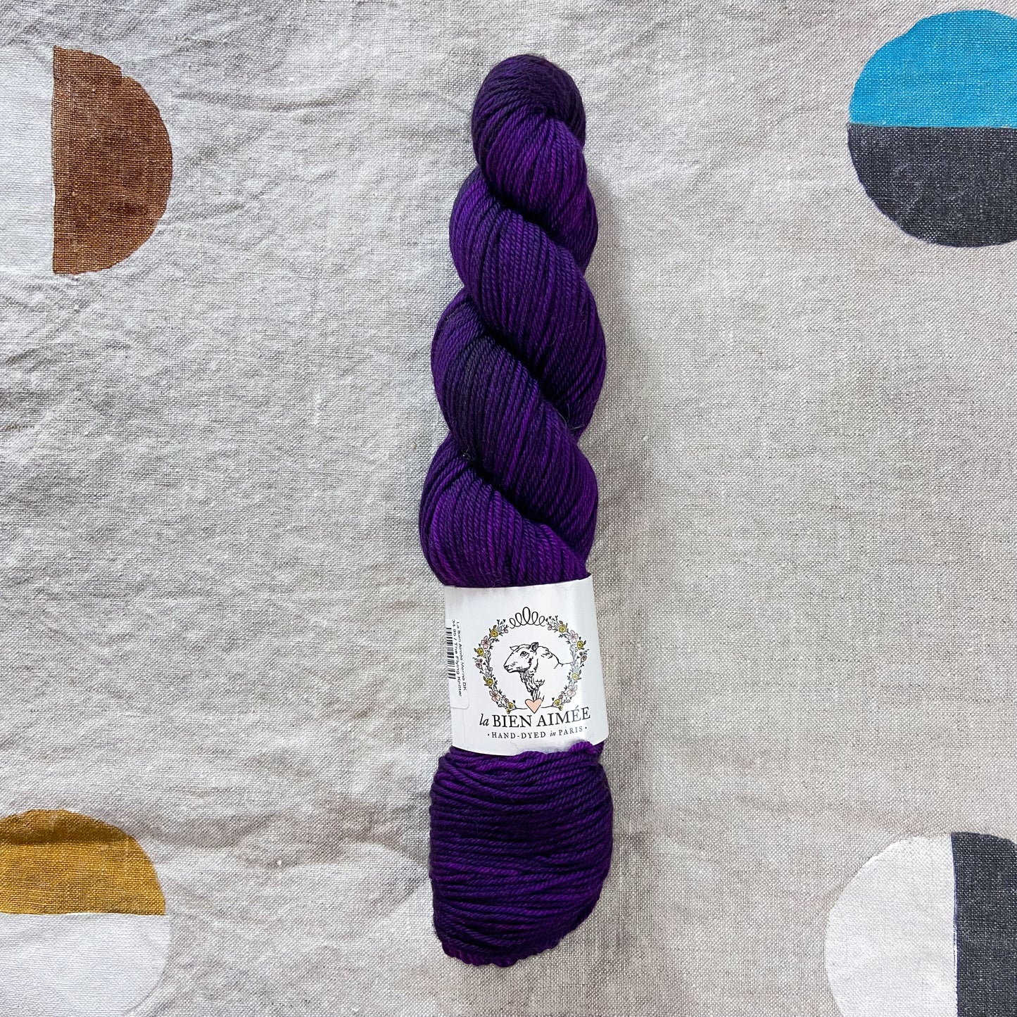 An image of a skein of DK weight yarn in a deep purple color with a white ‘La Bien Aimee’ label against a canvas background.