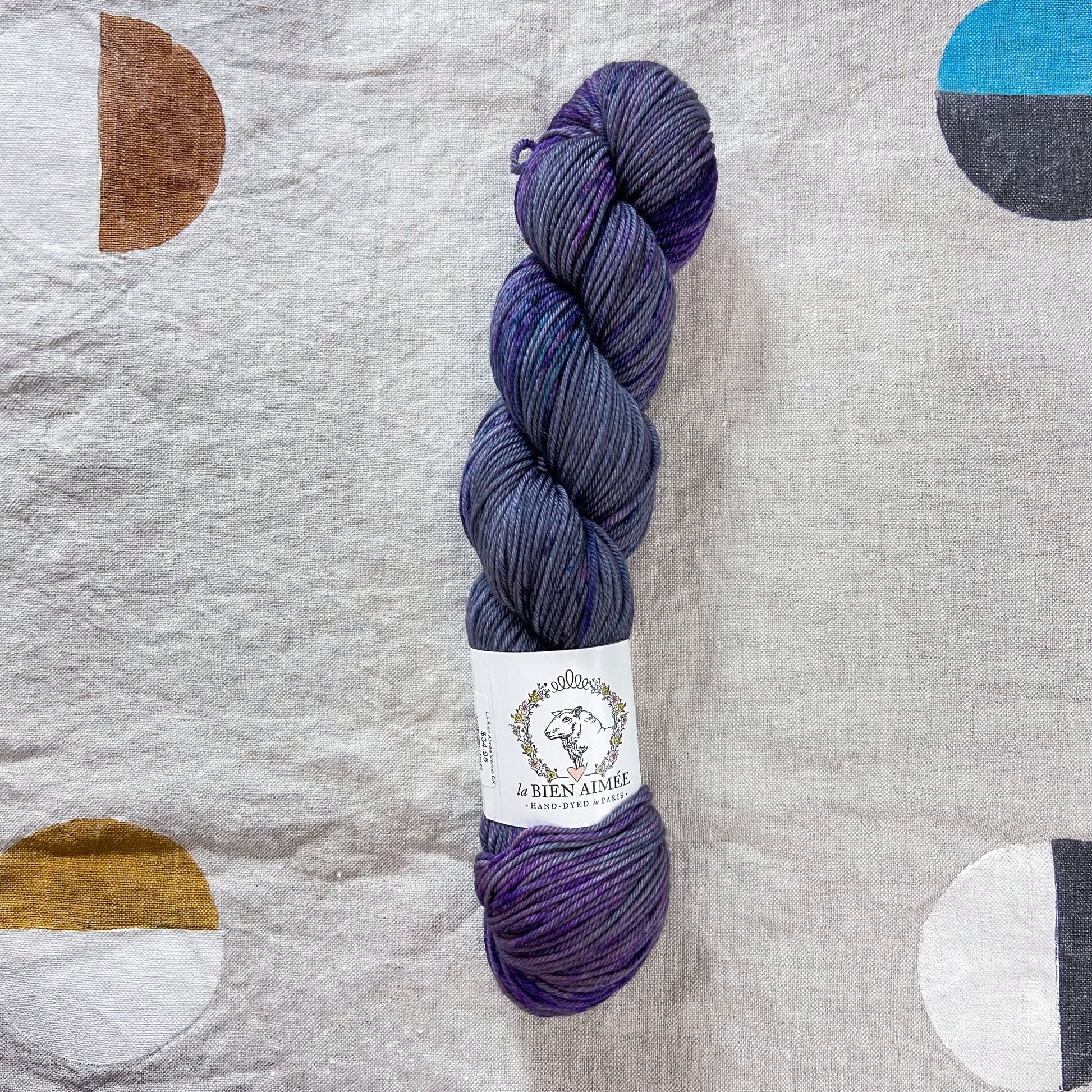 An image of a skein of DK weight yarn in shades of blue gray and purple with a white ‘La Bien Aimee’ label against a canvas background.