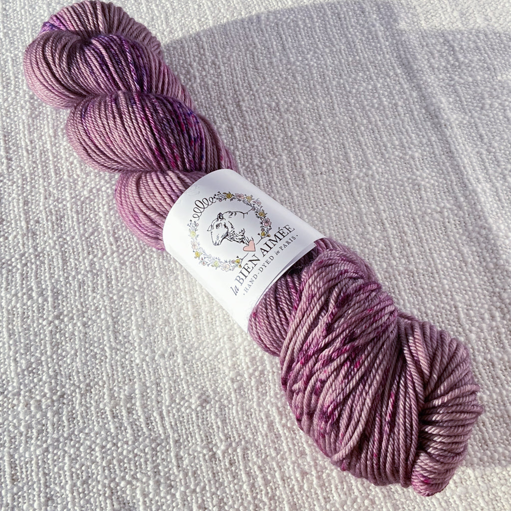 An image of a skein of DK weight yarn in a mauve color with speckles of purple and magenta, with a white ‘La Bien Aimee’ label against a canvas background.