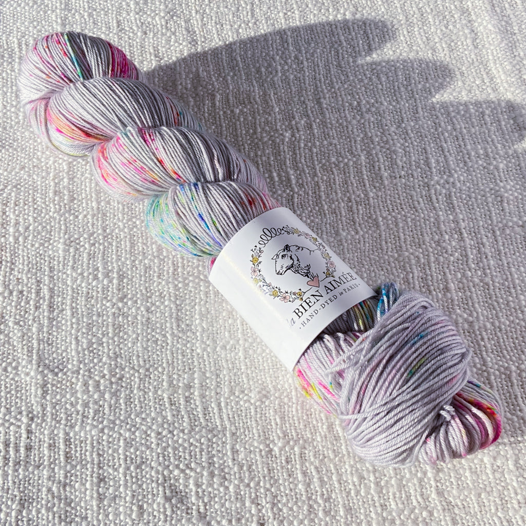 An image of a skein of sock yarn in a gray base with speckles of blue, green, and pink, and a white ‘La Bien Aimee’ label against a canvas background.