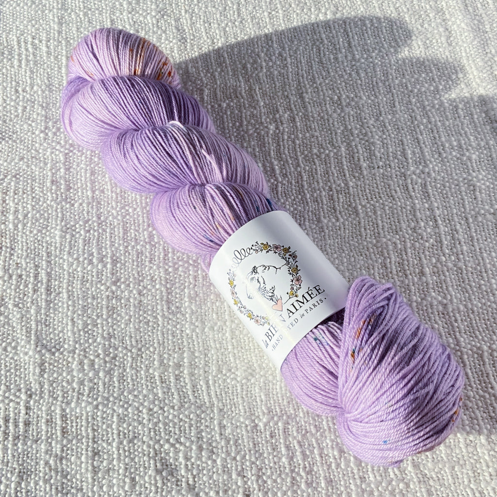 An image of a skein of sock yarn in a light purple base with speckles of orange and pink and a white ‘La Bien Aimee’ label against a canvas background.