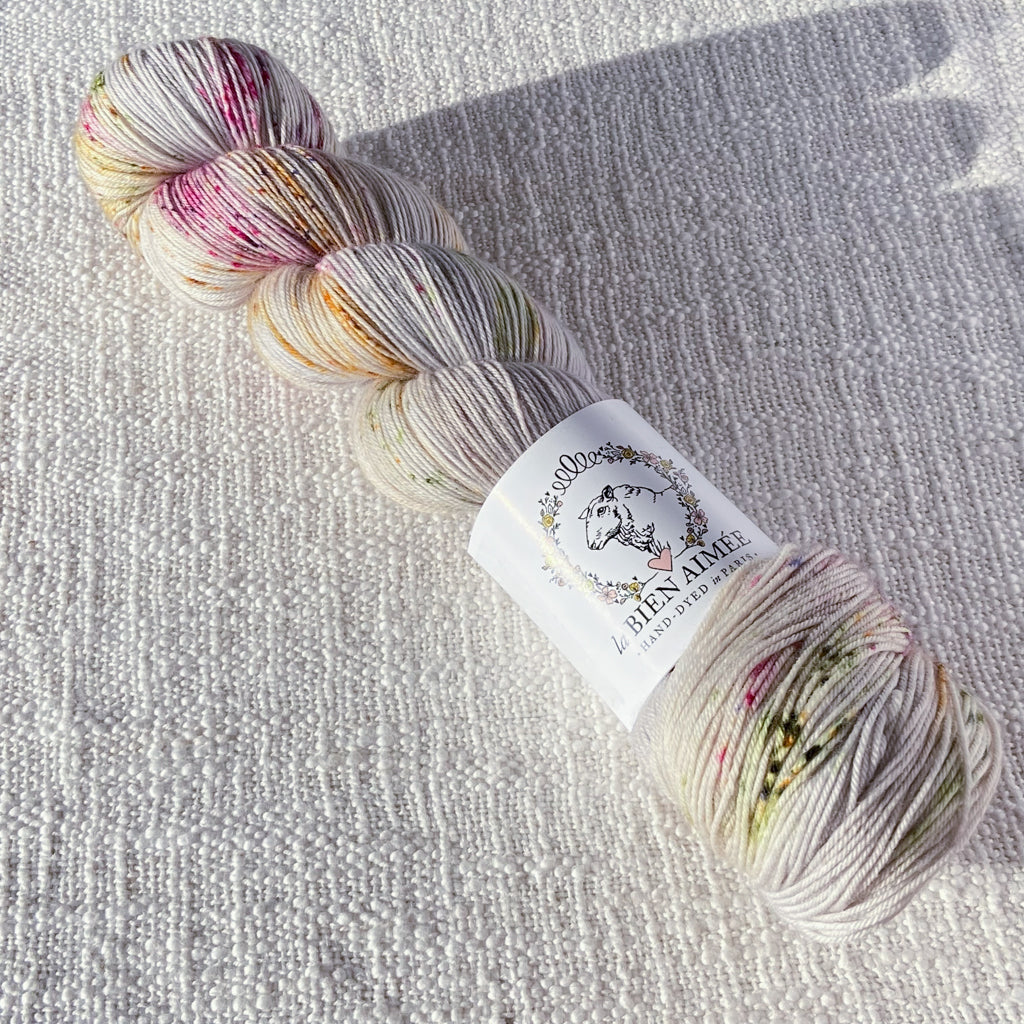 An image of a skein of sock yarn in a cream base with speckles of pink, orange, and dark green, with a white ‘La Bien Aimee’ label against a canvas background.